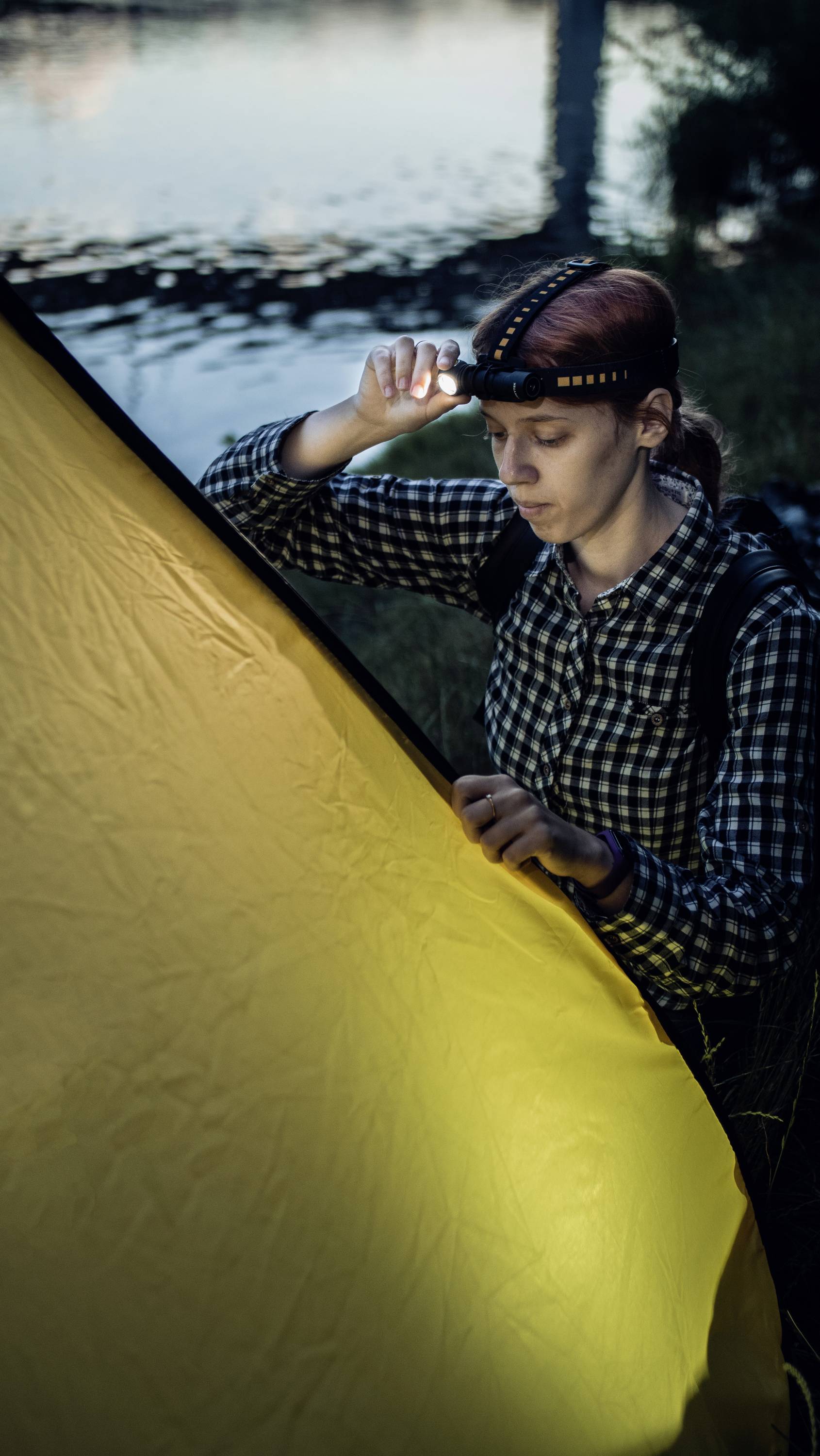 A person stands beside a yellow tent on the lakeshore, wearing a head torch. It is evening, and the torch is illuminating the scene.