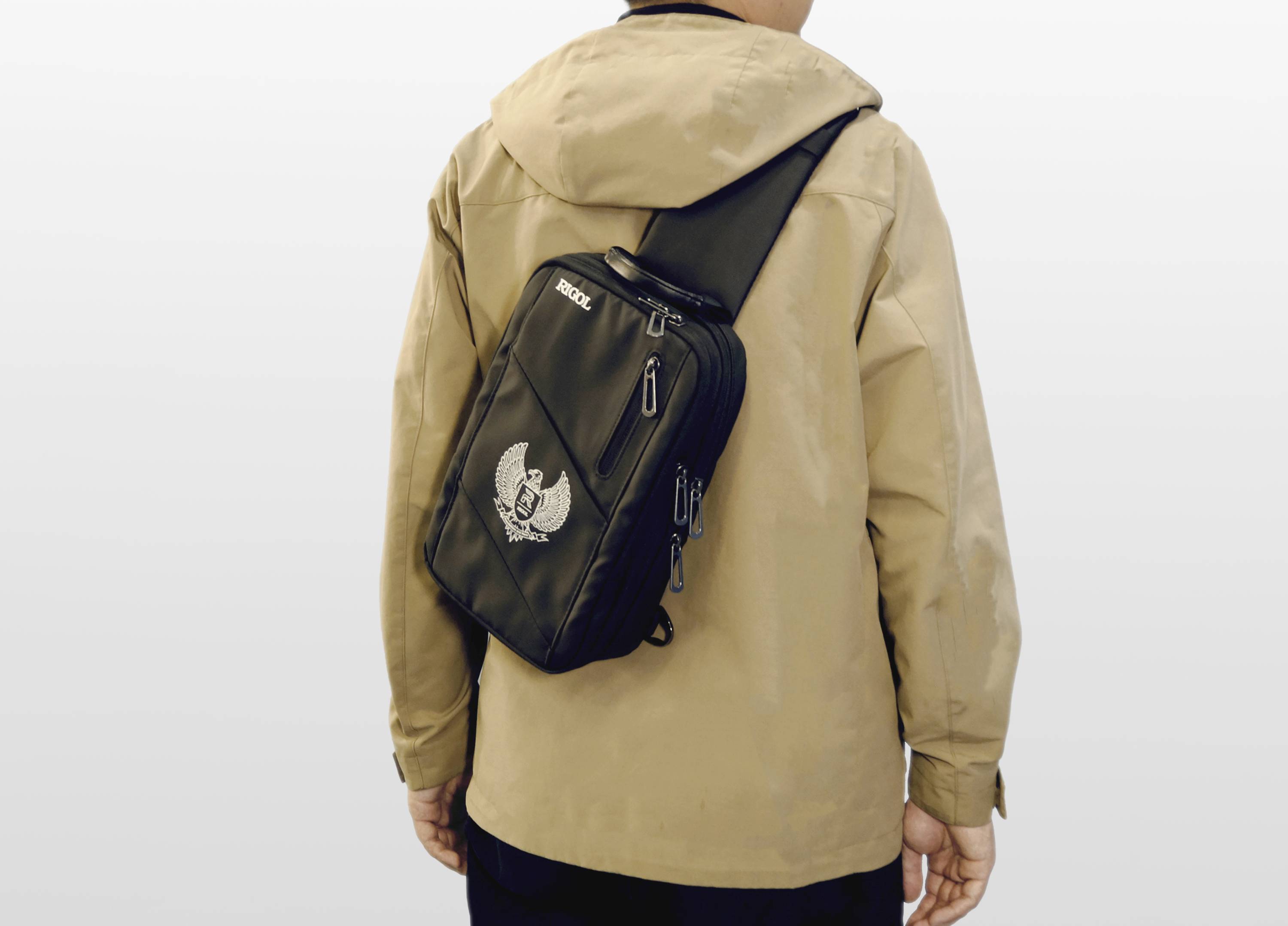 A person wearing a beige jacket is carrying a black shoulder bag with an emblem on the back.