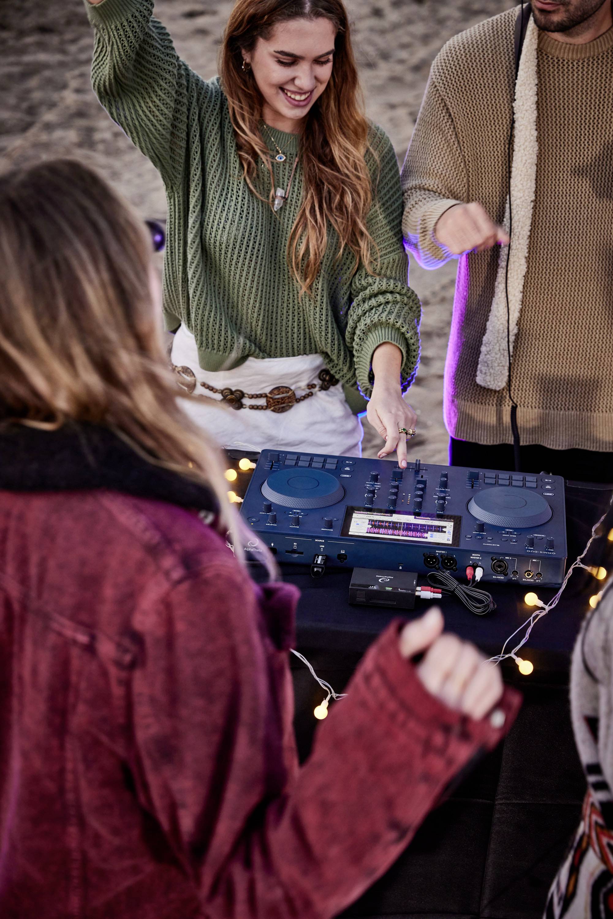 A group of young adults are dancing merrily around a DJ booth on the beach. Fairy lights decorate the table. The atmosphere is exuberant.