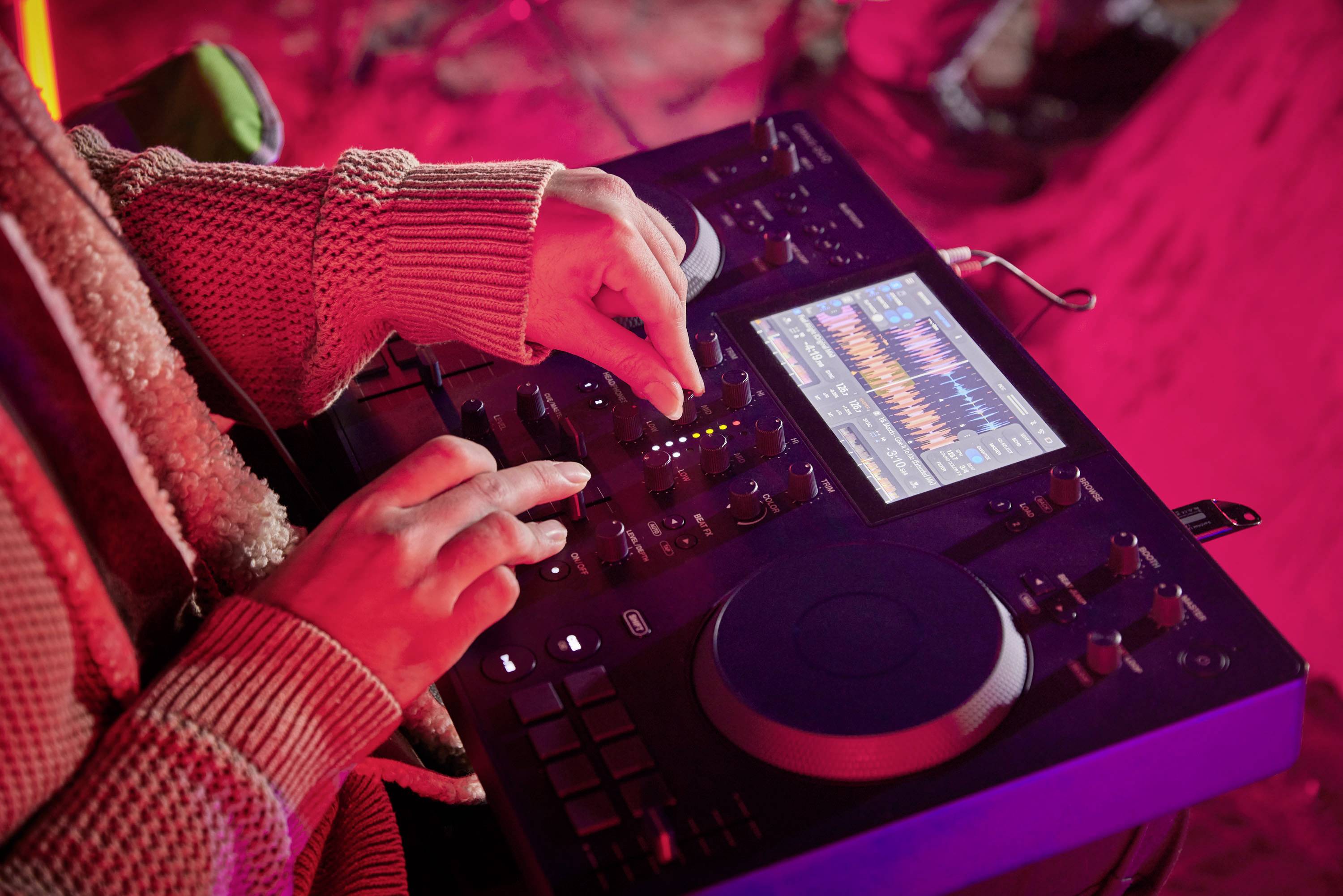 A person is operating a DJ mixing desk with a display showing music data and waveforms. The environment is illuminated with red lighting.