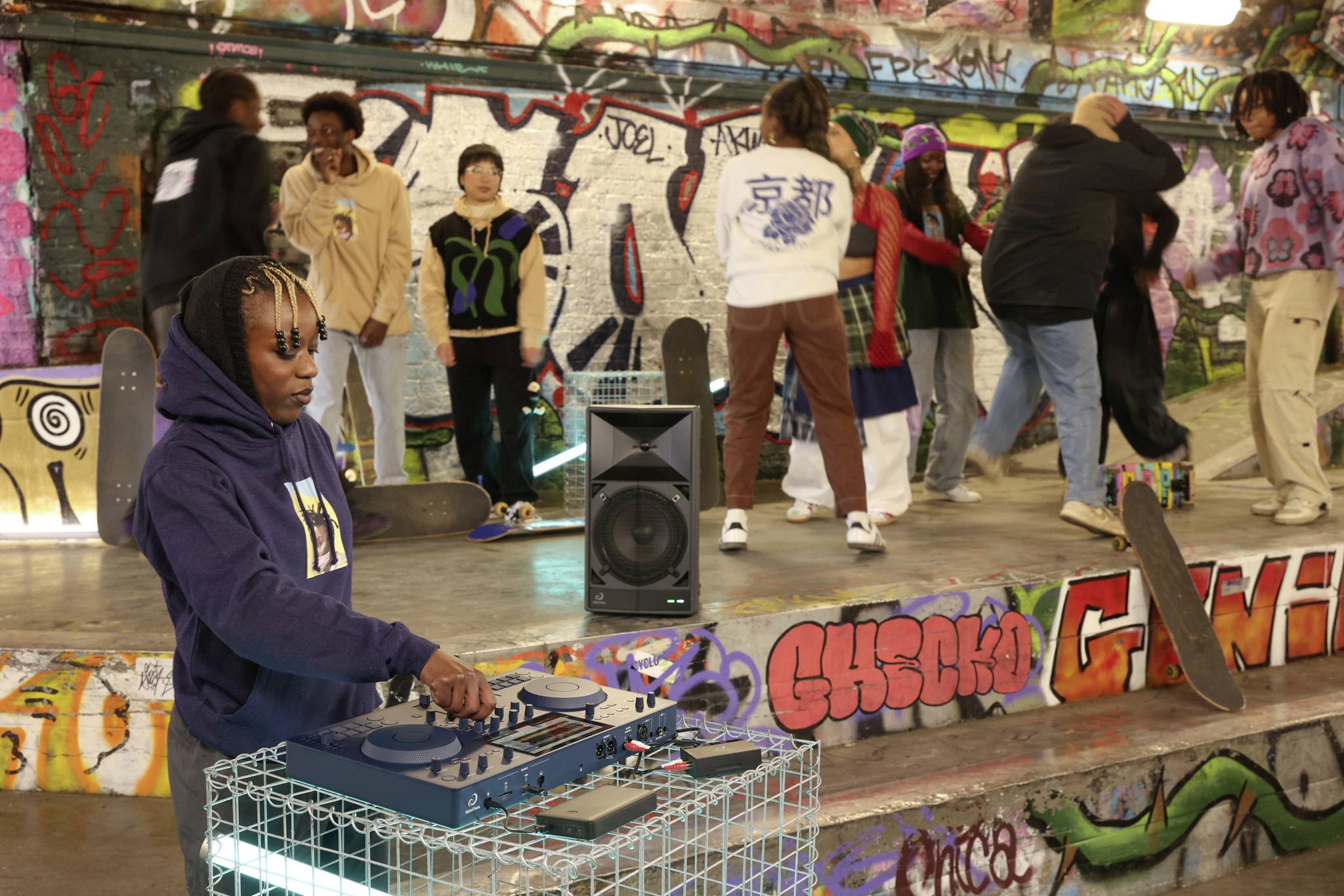 A person in a hoodie mixing music at a DJ booth in a graffiti-sprayed room. Young people in the background are dancing and skating.