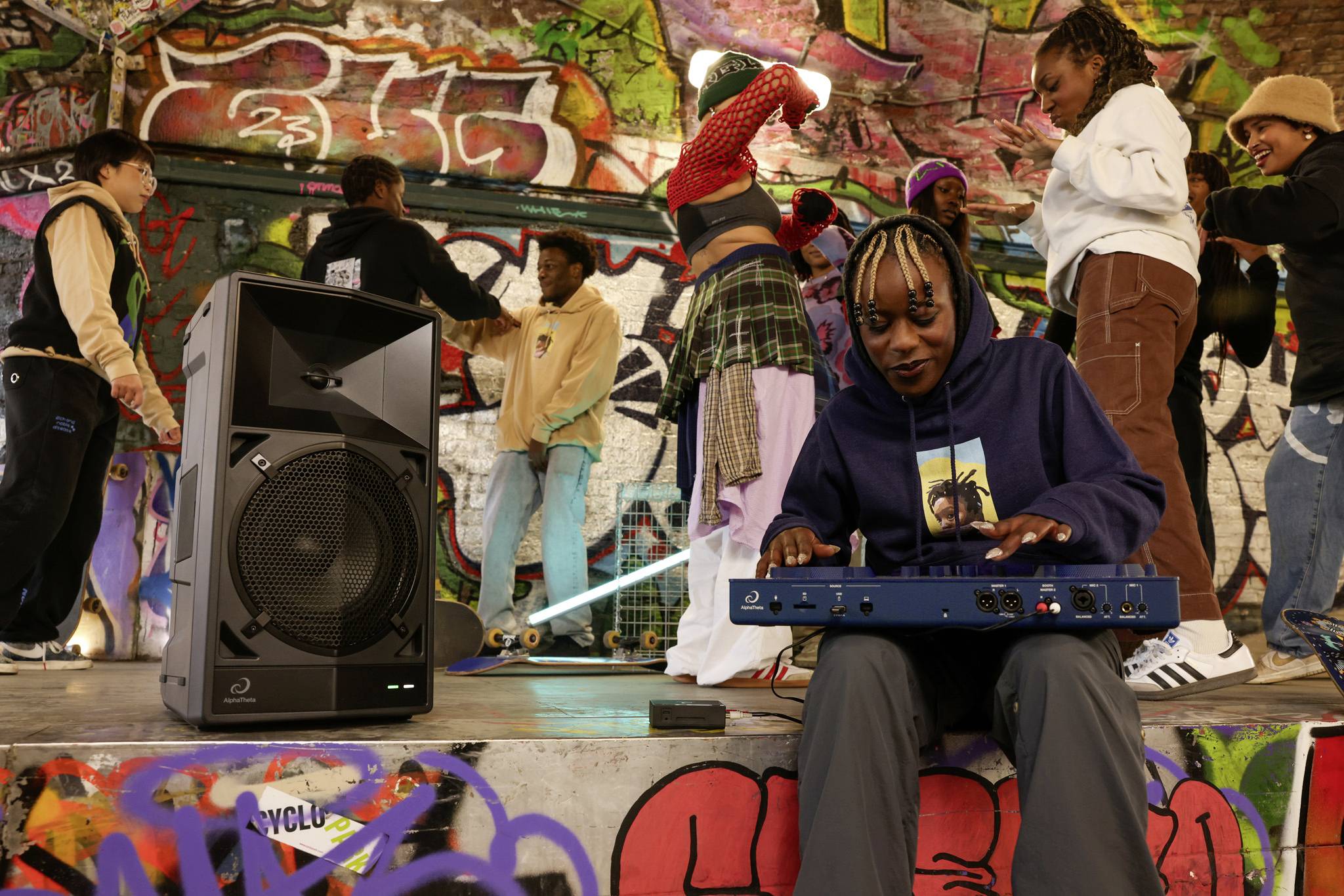 A person sits at a mixing desk playing music. In the background, people dance in colourful clothing in front of a graffiti-covered wall.