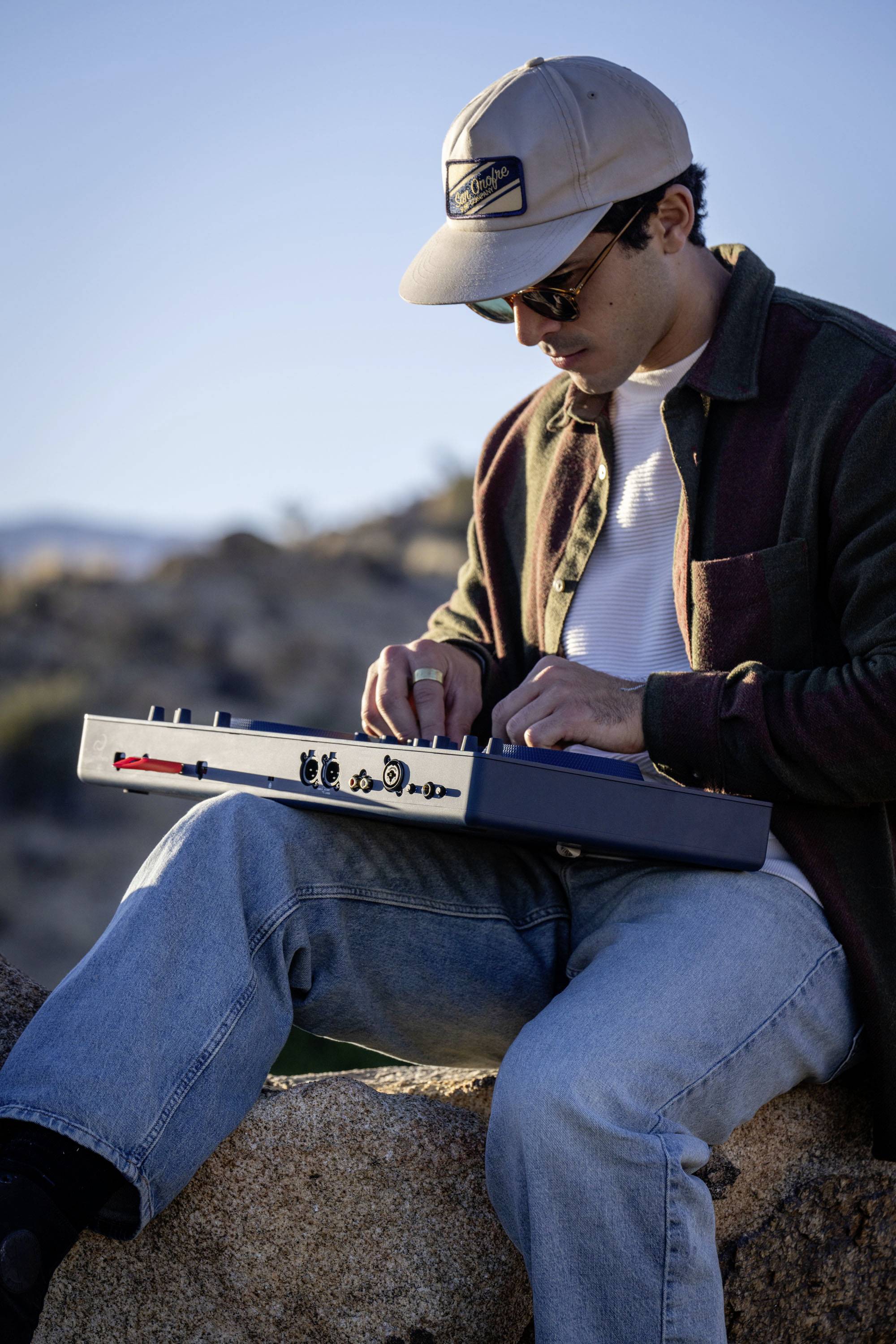 A man sits on a rock in a rural setting, playing a portable electronic musical instrument.