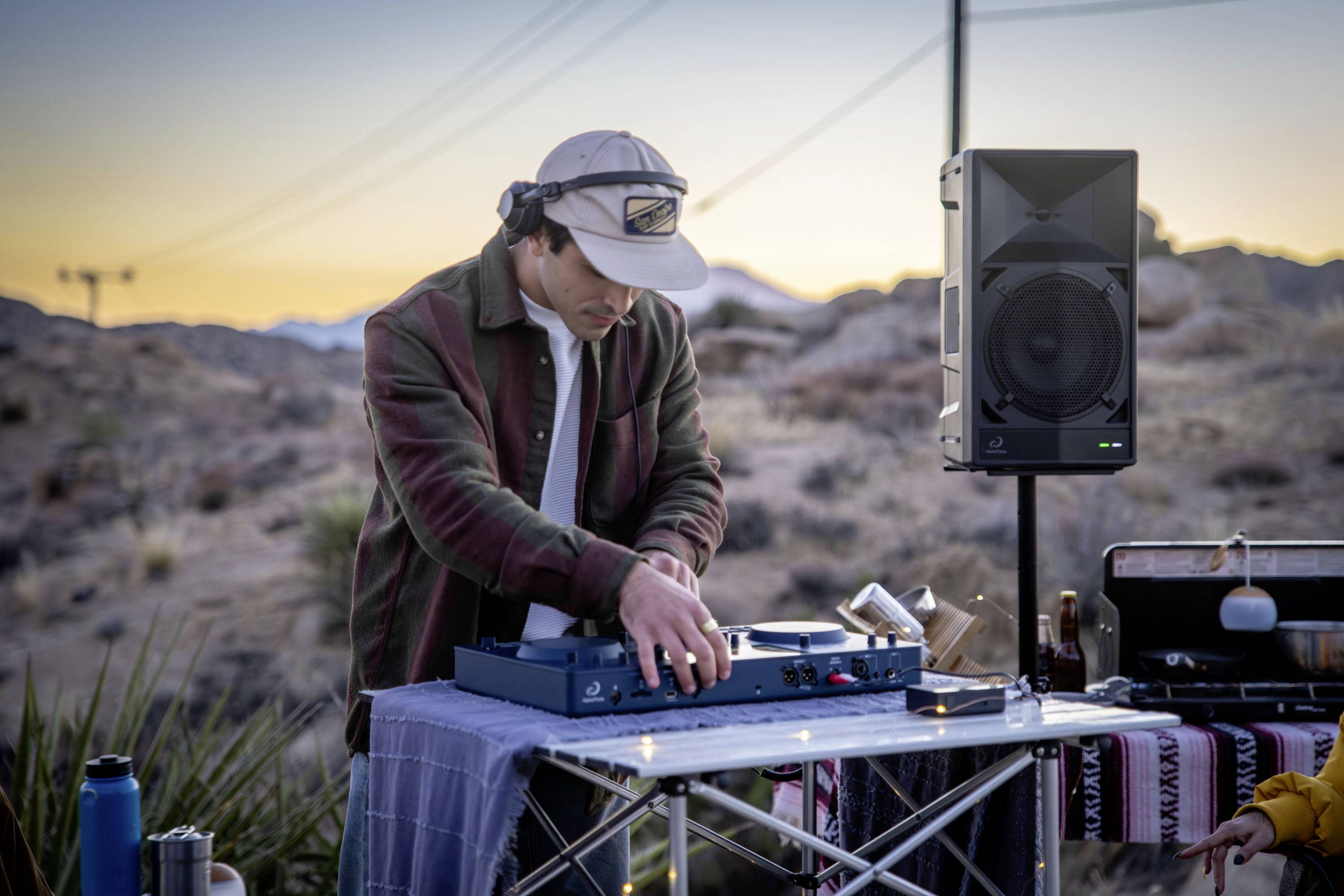 A DJ mixes music on a turntable in a desert landscape at sunset. A speaker box stands nearby.