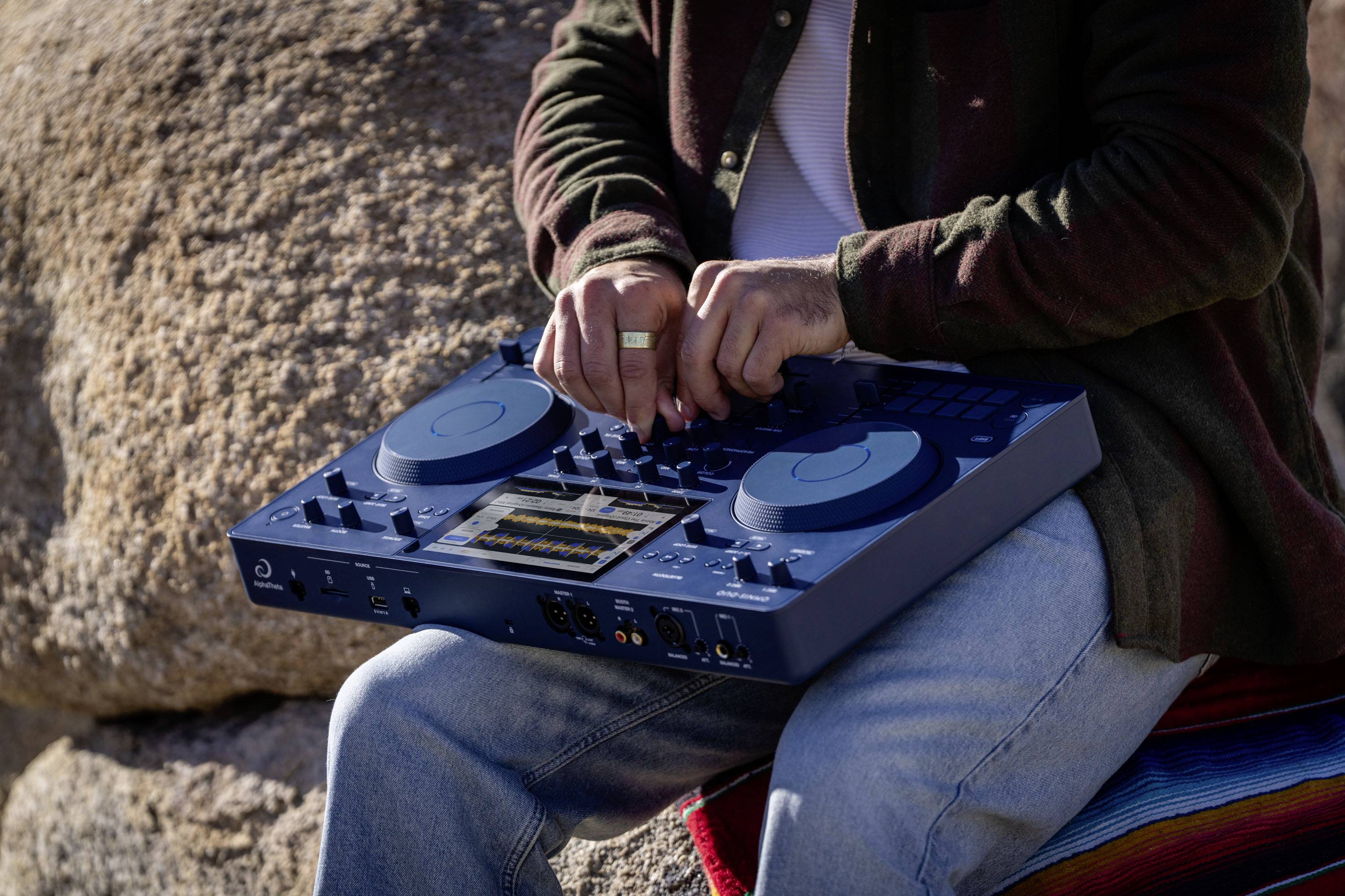 A person is operating a blue mixing desk outdoors, sitting on a multi-coloured striped cloth and surrounded by rocks.