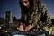 A woman with curly hair stands outdoors at dusk, mixing music on a DJ controller. Speakers in the background.
