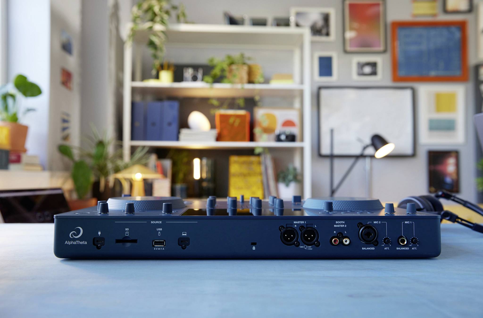 A music mixing desk on a table, with a bookshelf in the background and pictures and plants on the wall.