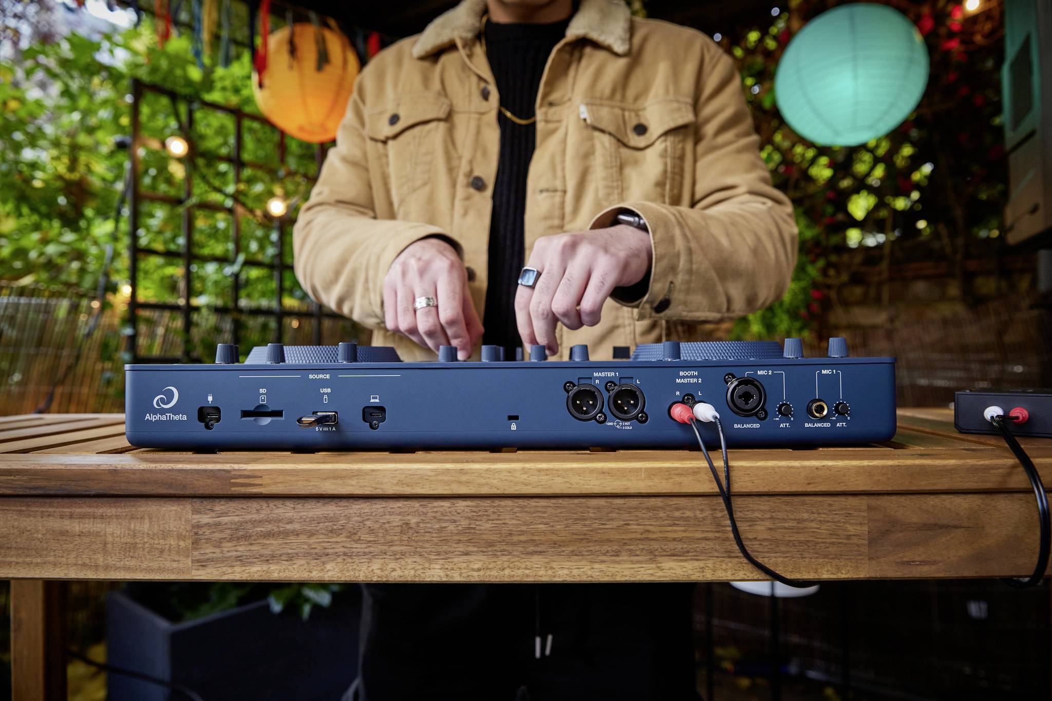 A person is operating a DJ mixing desk outdoors. Various colourful lights and plants are visible in the background.