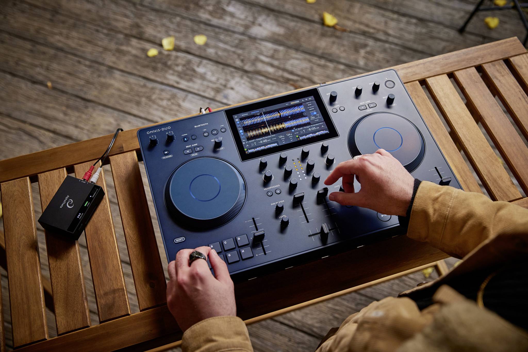 A person is mixing music with a DJ controller on a wooden table. A small audio device is connected beside it.