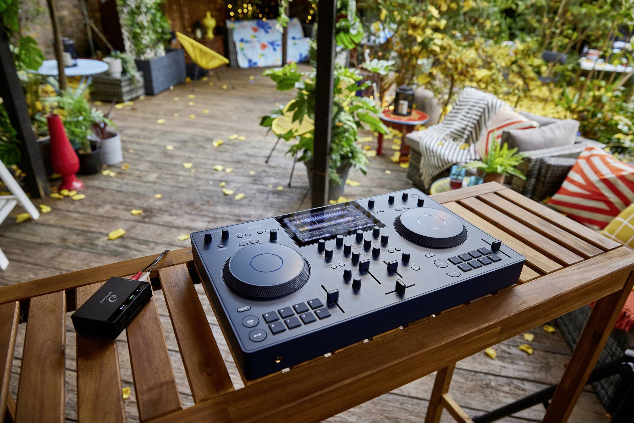 DJ controller on a wooden table on a terrace with colourful cushions and plants in the background.