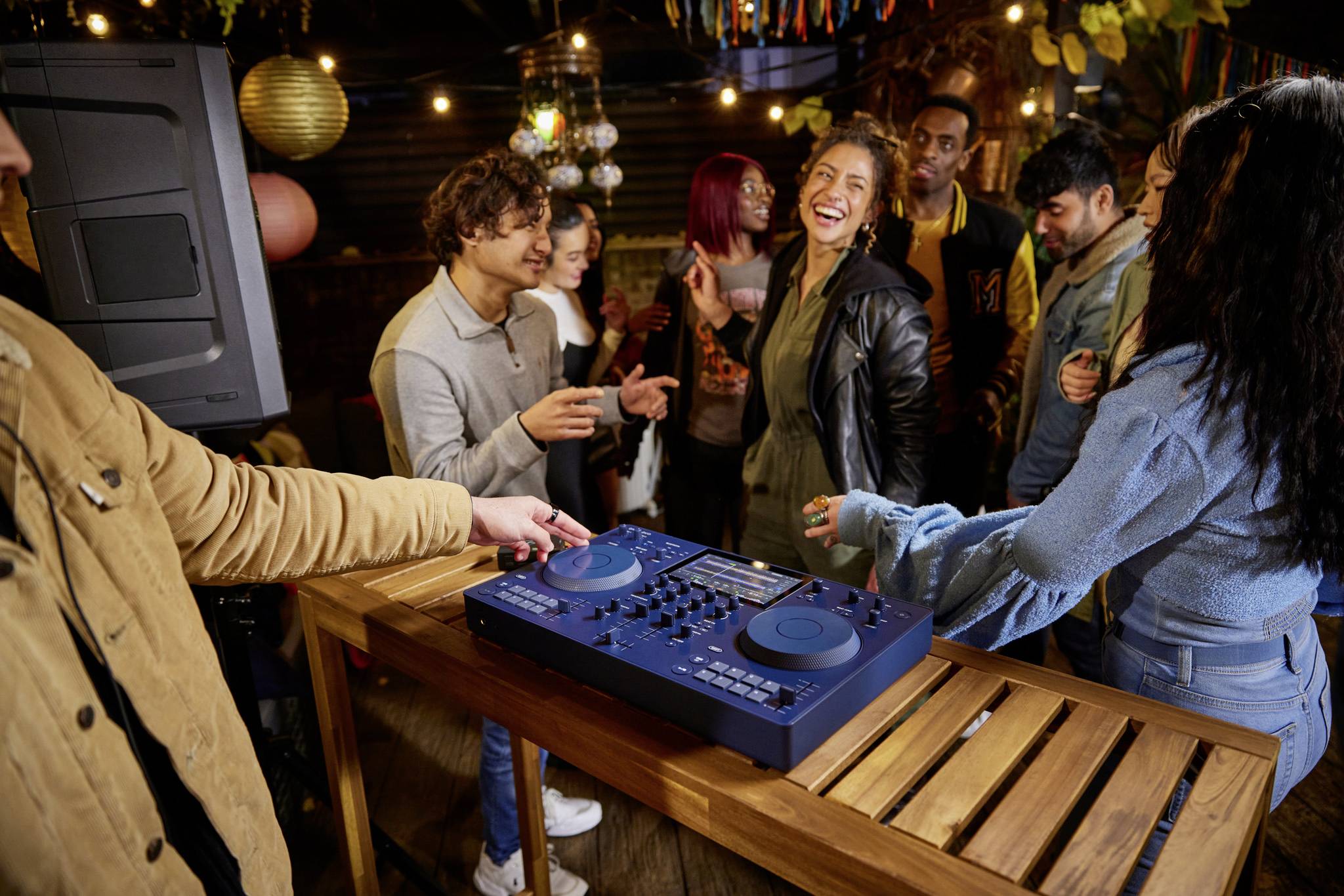 A group of people are dancing merrily around a DJ booth at a party. Lights and decorations in the background create a festive atmosphere.