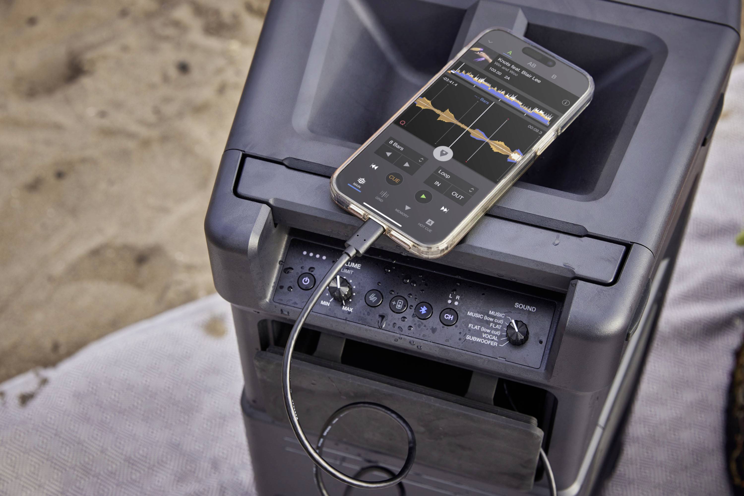 A smartphone connected to a speaker is displaying a music app. The speaker is positioned on a sandy surface.
