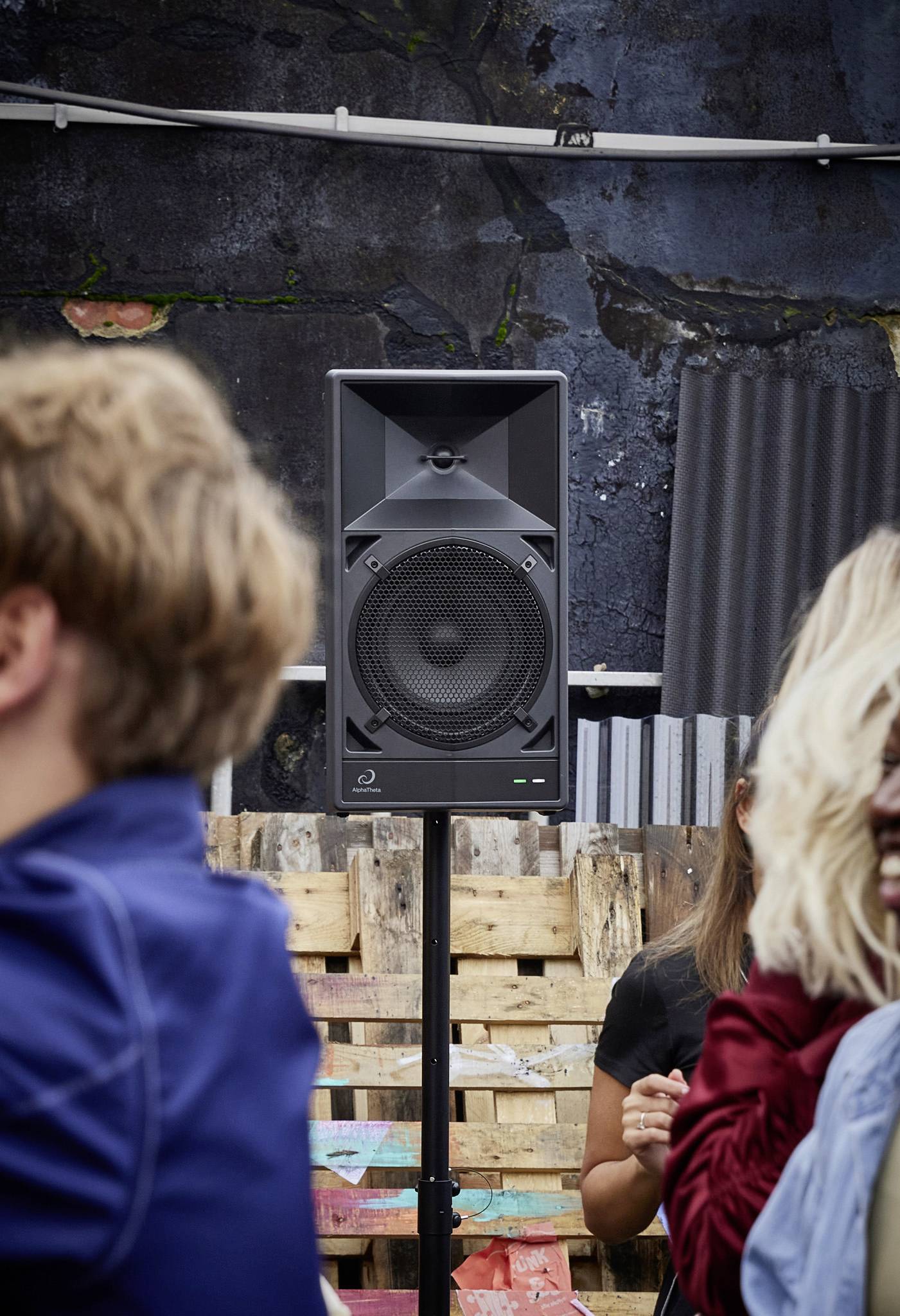 Speakers on a stand outdoors at an event. People are standing nearby, with pallets and a wall in the background.