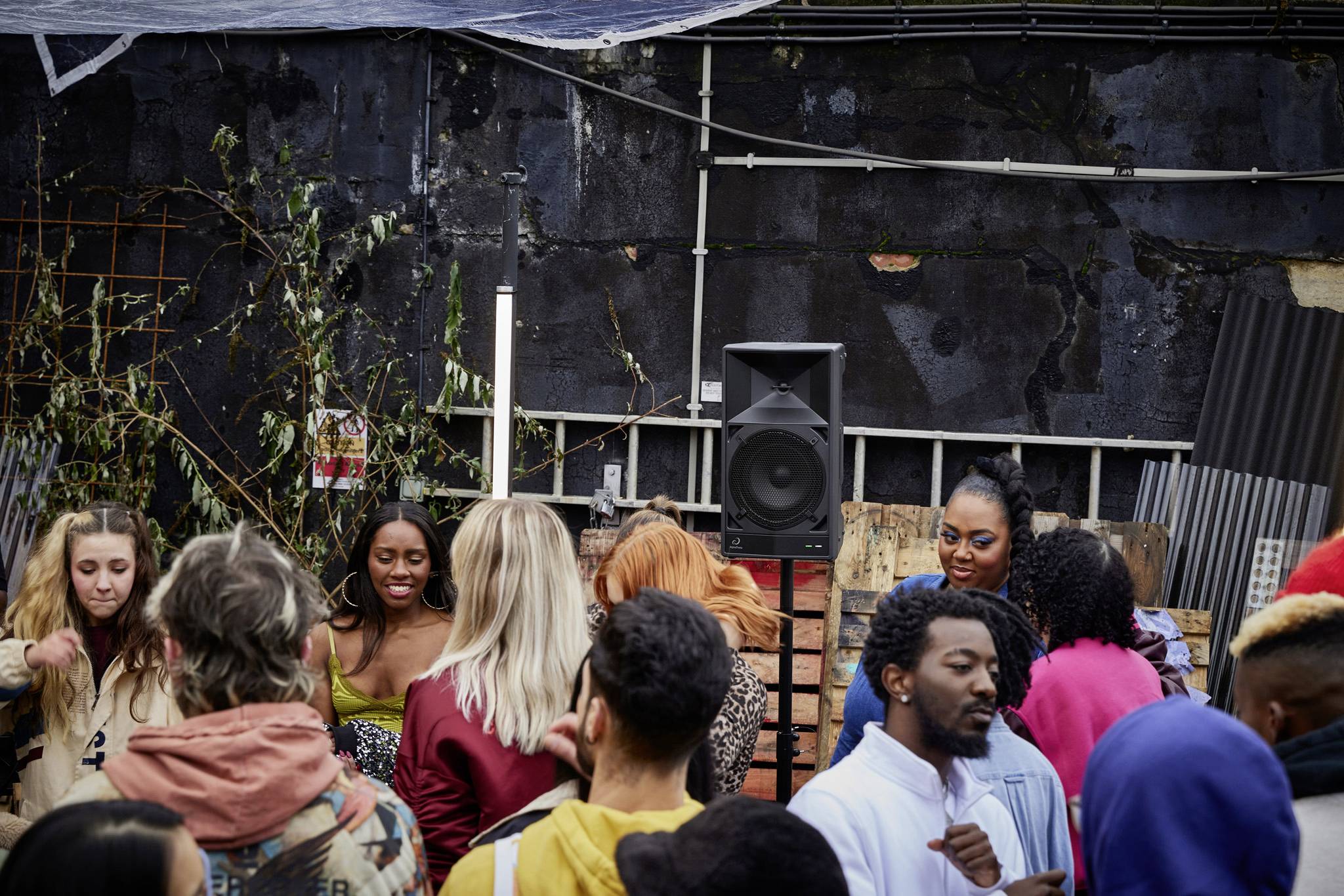 People are dancing joyfully in an outdoor setting. A group of people in colourful clothing can be seen. A speaker is visible in the background.