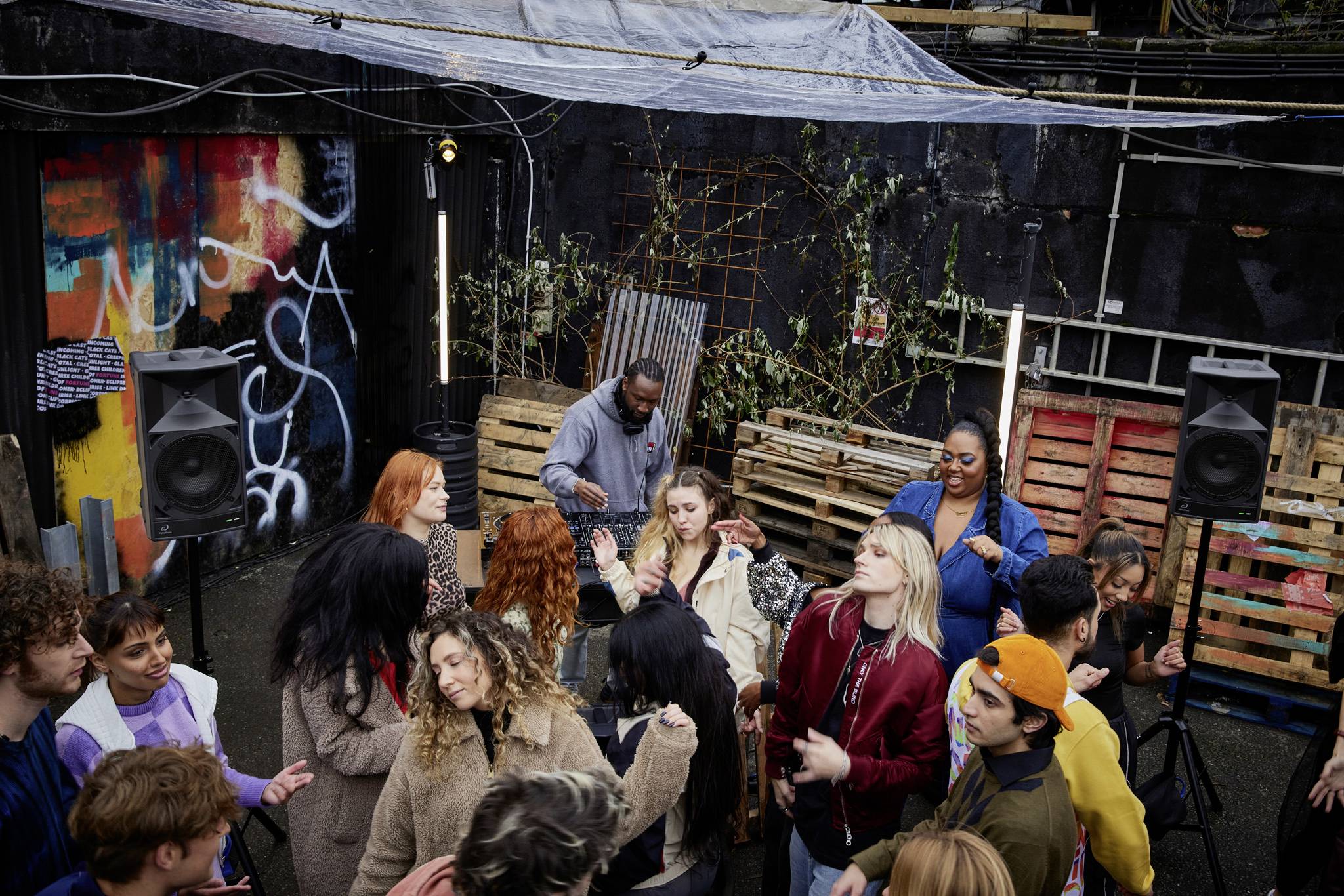 A group of people are dancing and chatting in an outdoor area with colourful wall art and pallet furniture. Relaxed, sociable atmosphere.