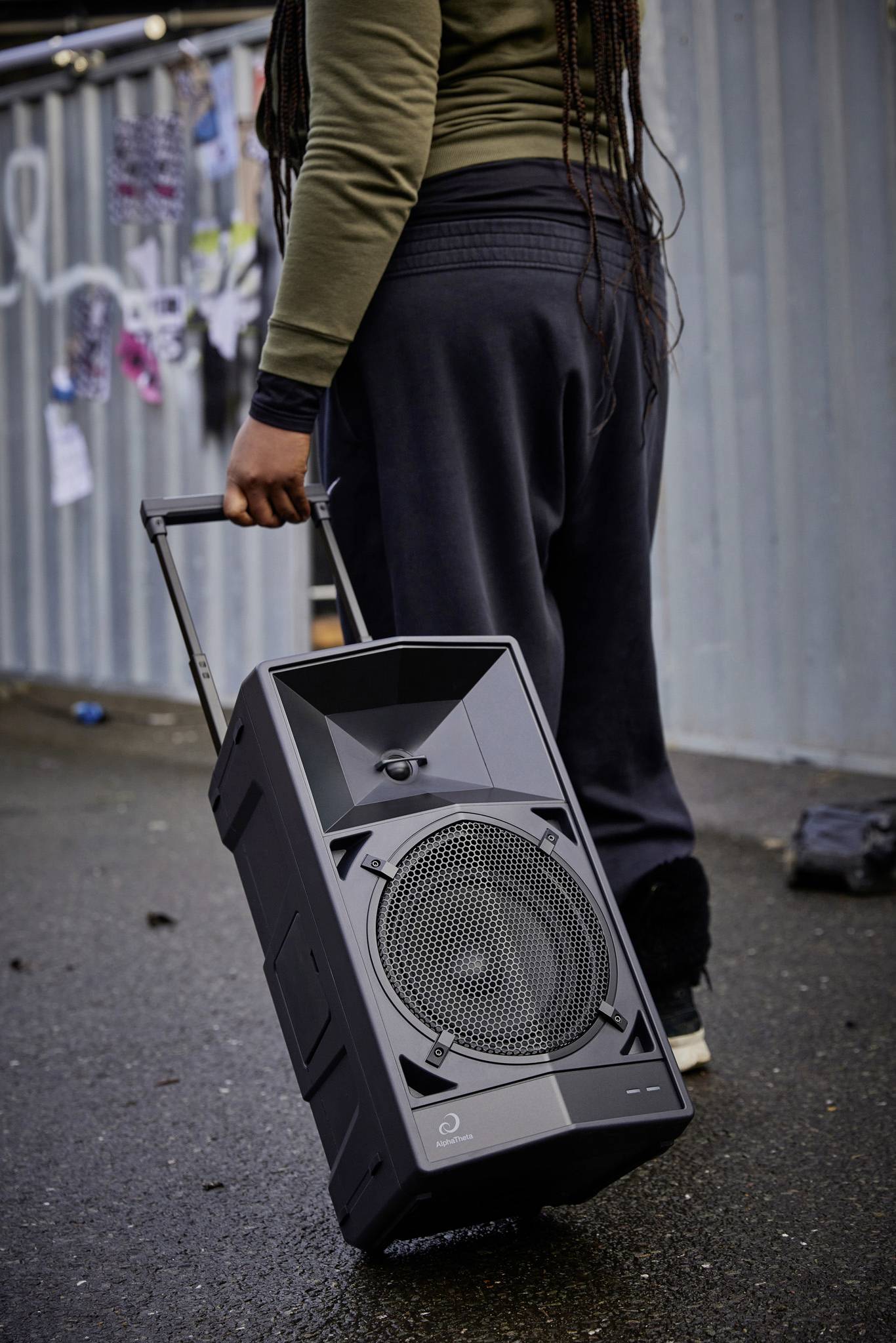 A person is carrying a portable speaker on wheels. In the background, various colourful garments are hanging to dry.