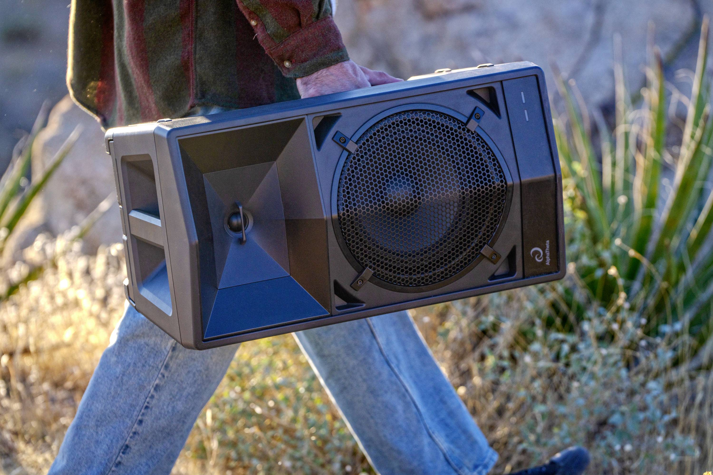 A person is carrying a large portable speaker through a dry, grassy landscape.