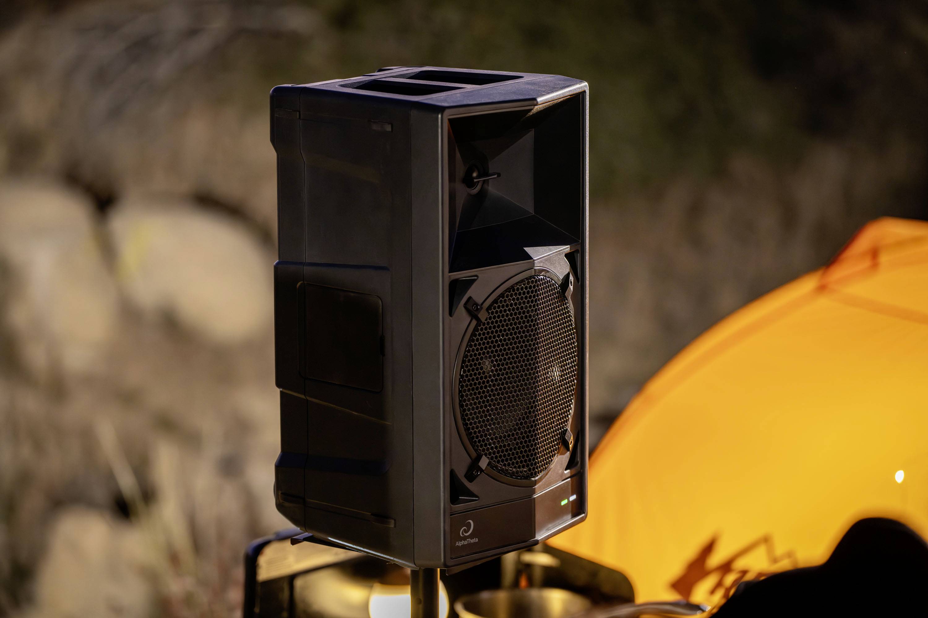 A single black speaker stands outdoors on a stand. Blurred rocks and an orange structure are visible in the background.