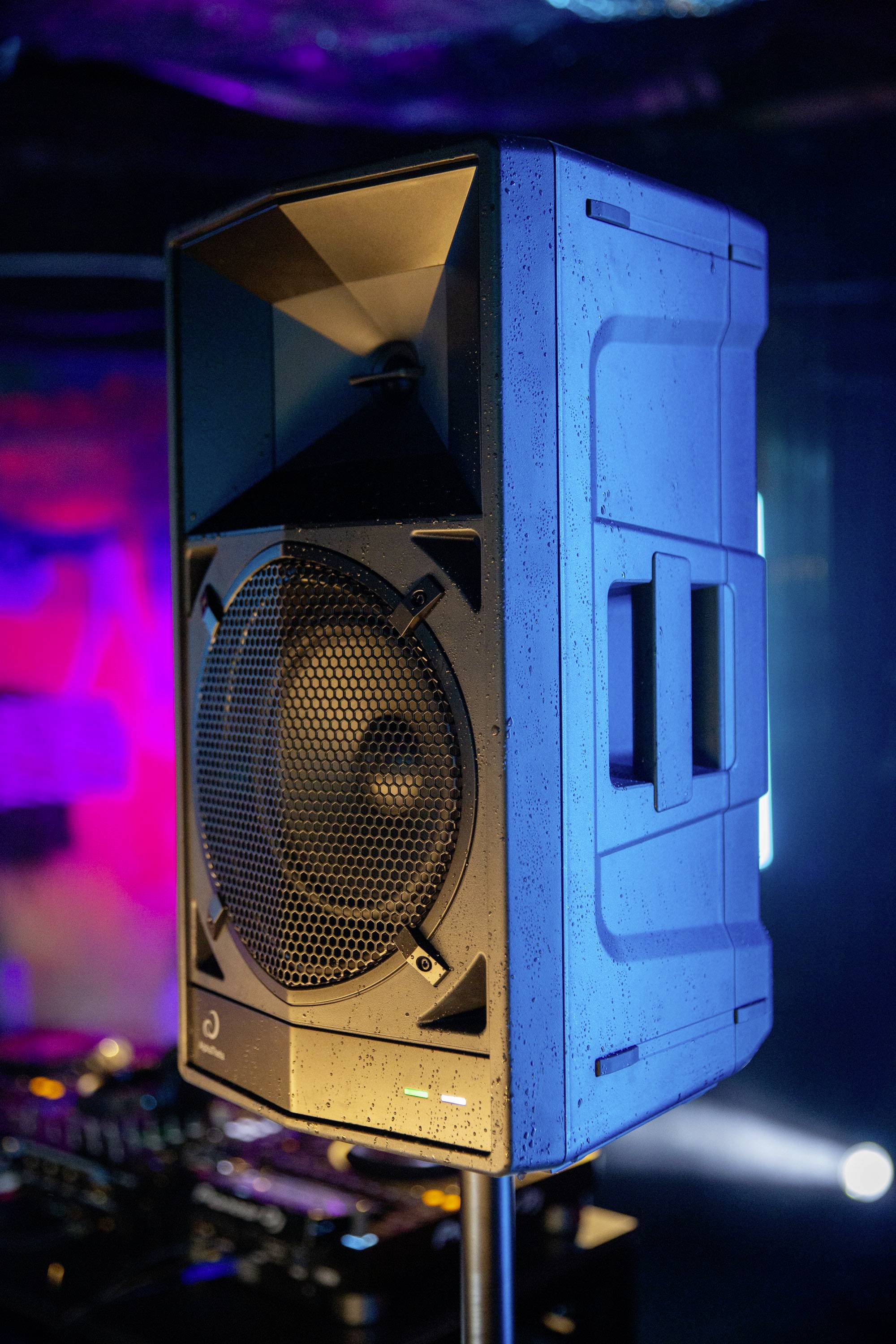 A large, standing speaker in a lit room with DJ equipment in the background.