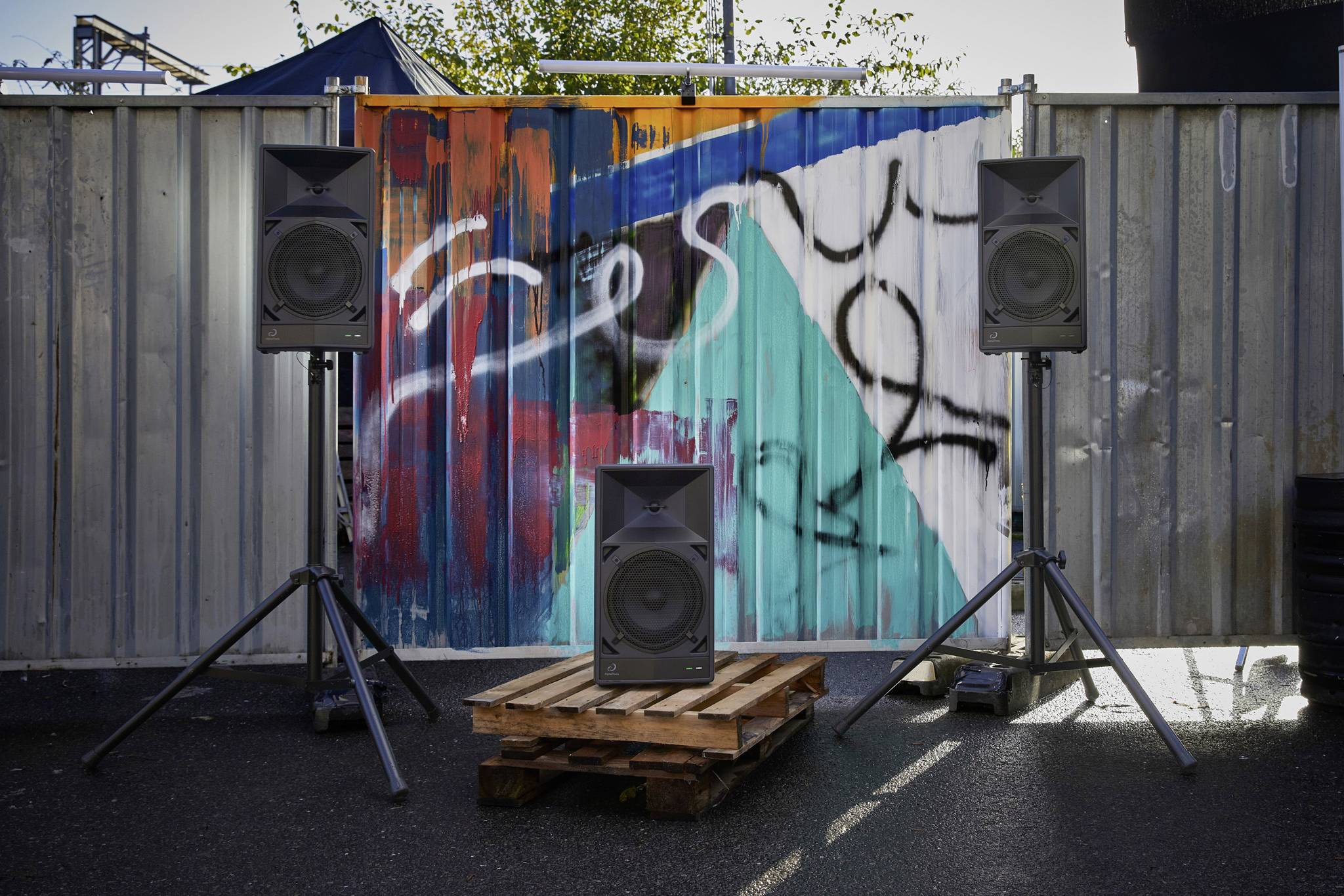 Three speakers stand on stands in front of a colourful, graffiti-painted metal wall. One speaker sits in the centre on a pallet.