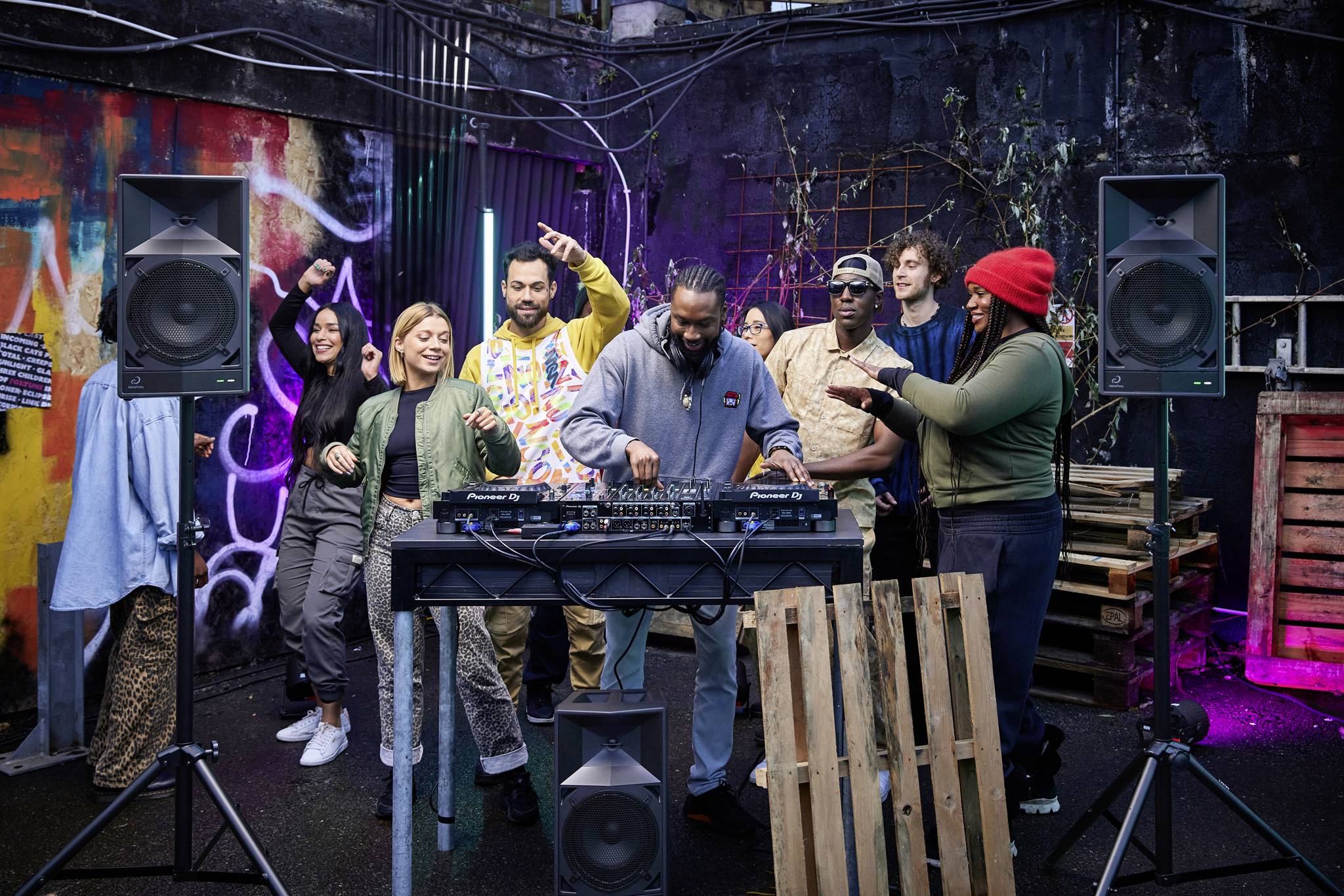 A group of people are dancing and interacting around a DJ who is playing music. The scene takes place outdoors in front of a vibrant wall decorated with graffiti.