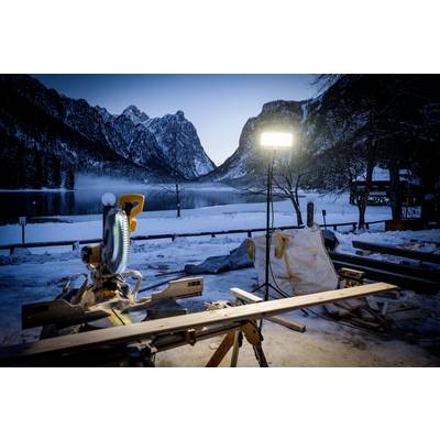 Wooden saw on a table in front of a snow-covered mountain lake at dusk, illuminated by a bright work light. Majestic mountain landscape in the background.