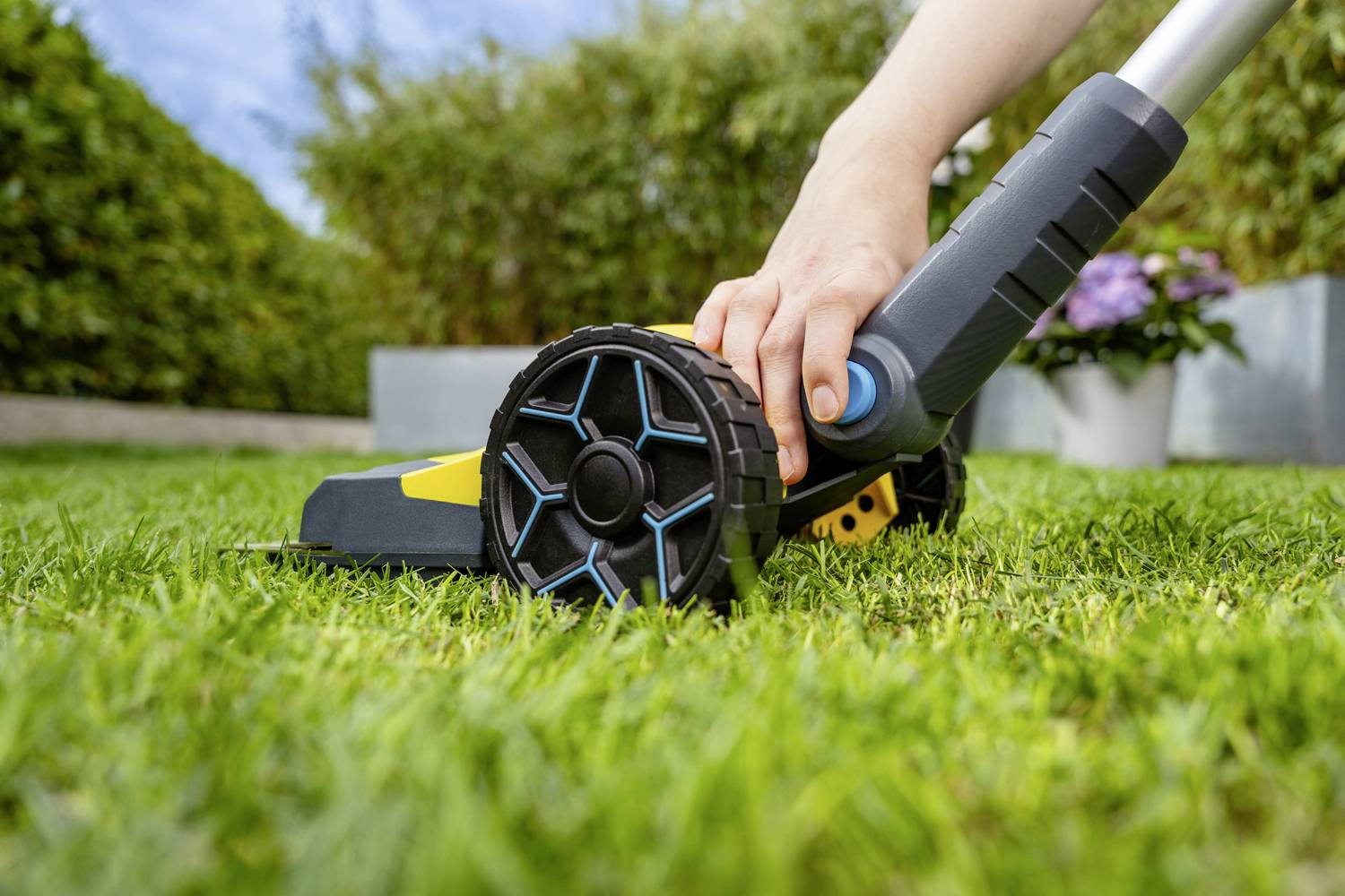 A hand is holding the handle of a lawnmower being pushed across a green lawn. Bushes and a flower are visible in the background.