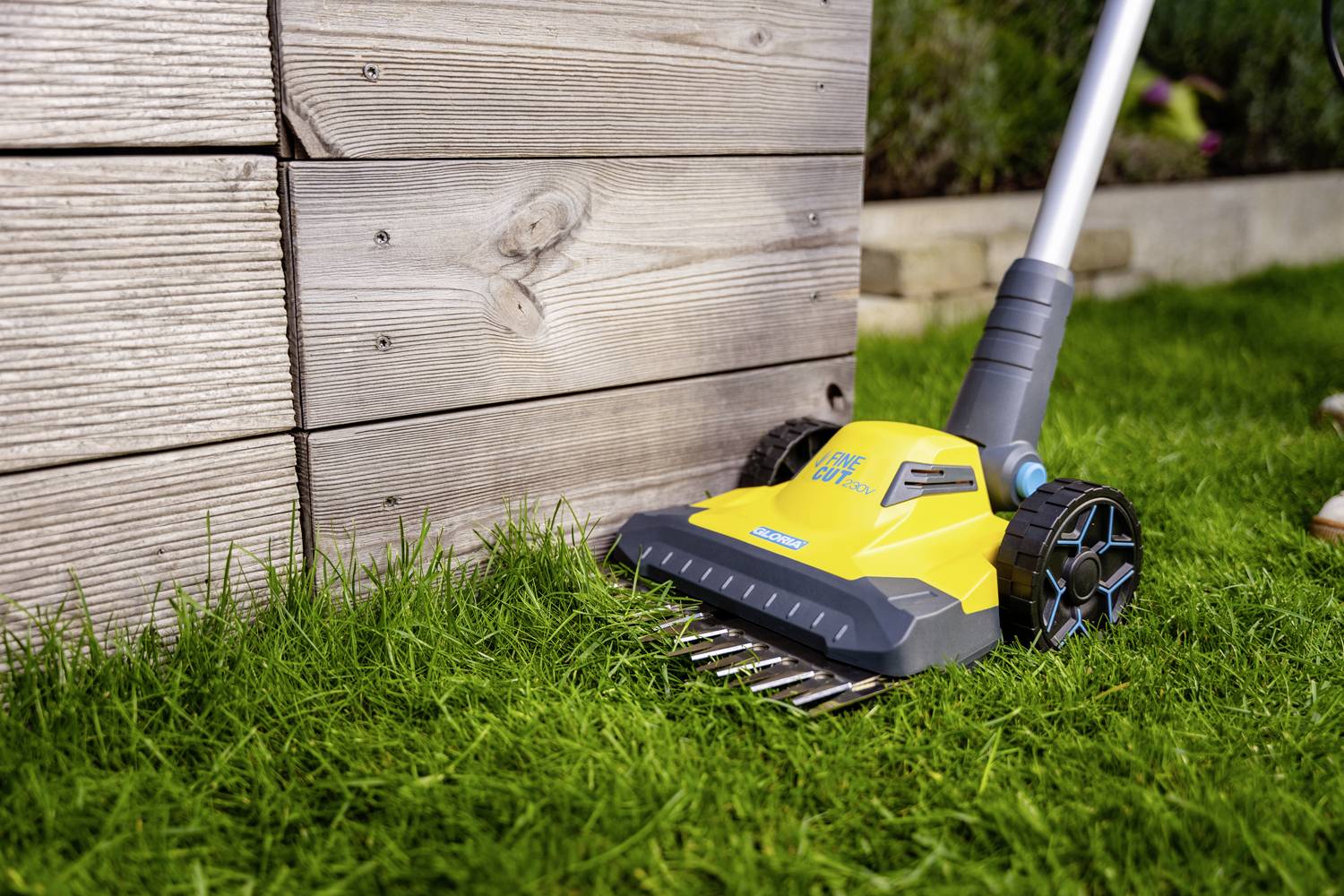 A yellow electric lawn edger is cutting grass along a wooden wall. It is positioned on a green lawn.