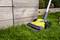 A yellow electric lawn edger is cutting grass along a wooden wall. It is positioned on a green lawn.