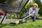 A man is using an electric lawn trimmer to cut the grass beneath a garden chair. Plants and flowers are visible in the background.