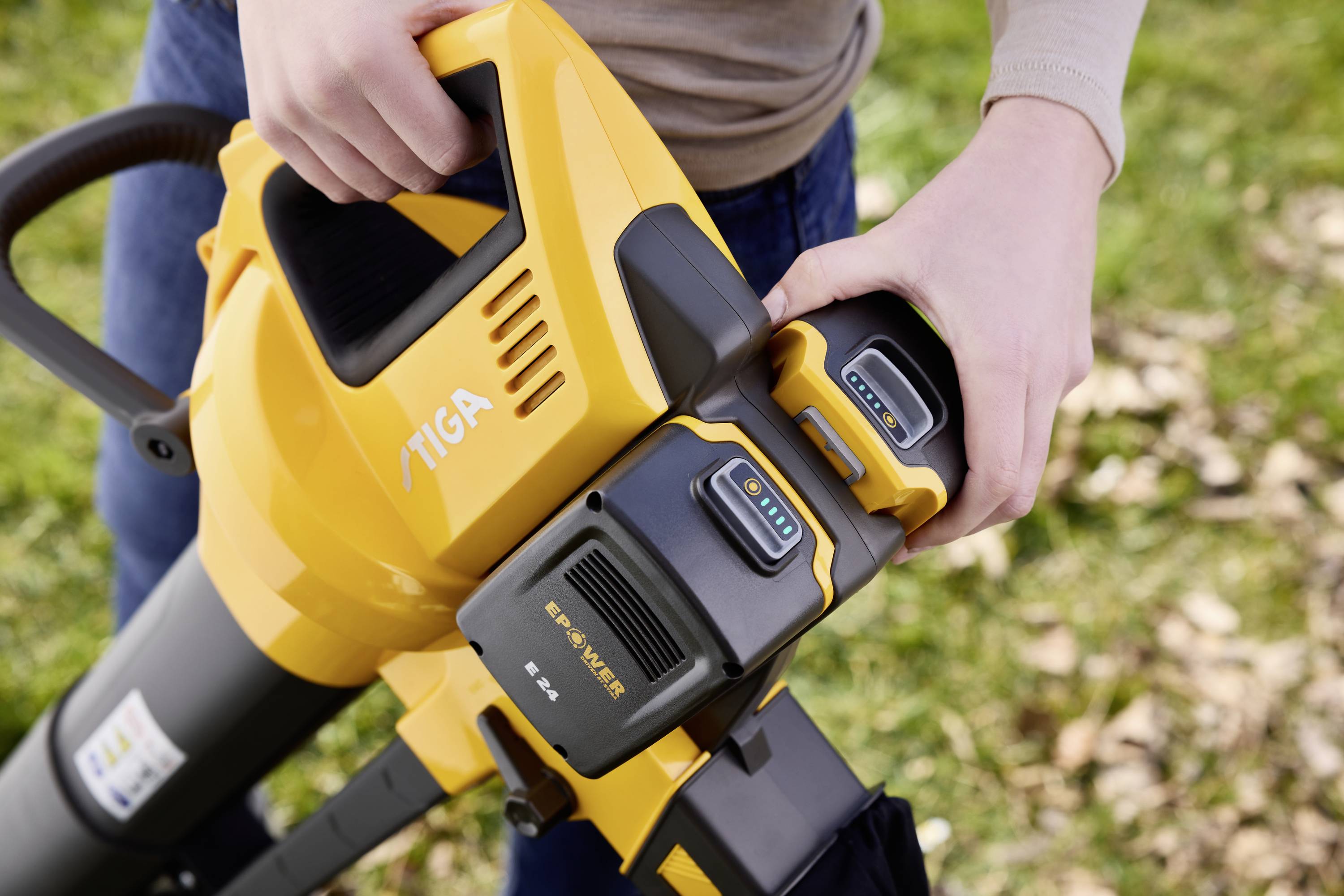 A person is changing the battery of a yellow leaf blower in a garden. The battery is displaying its charge status.