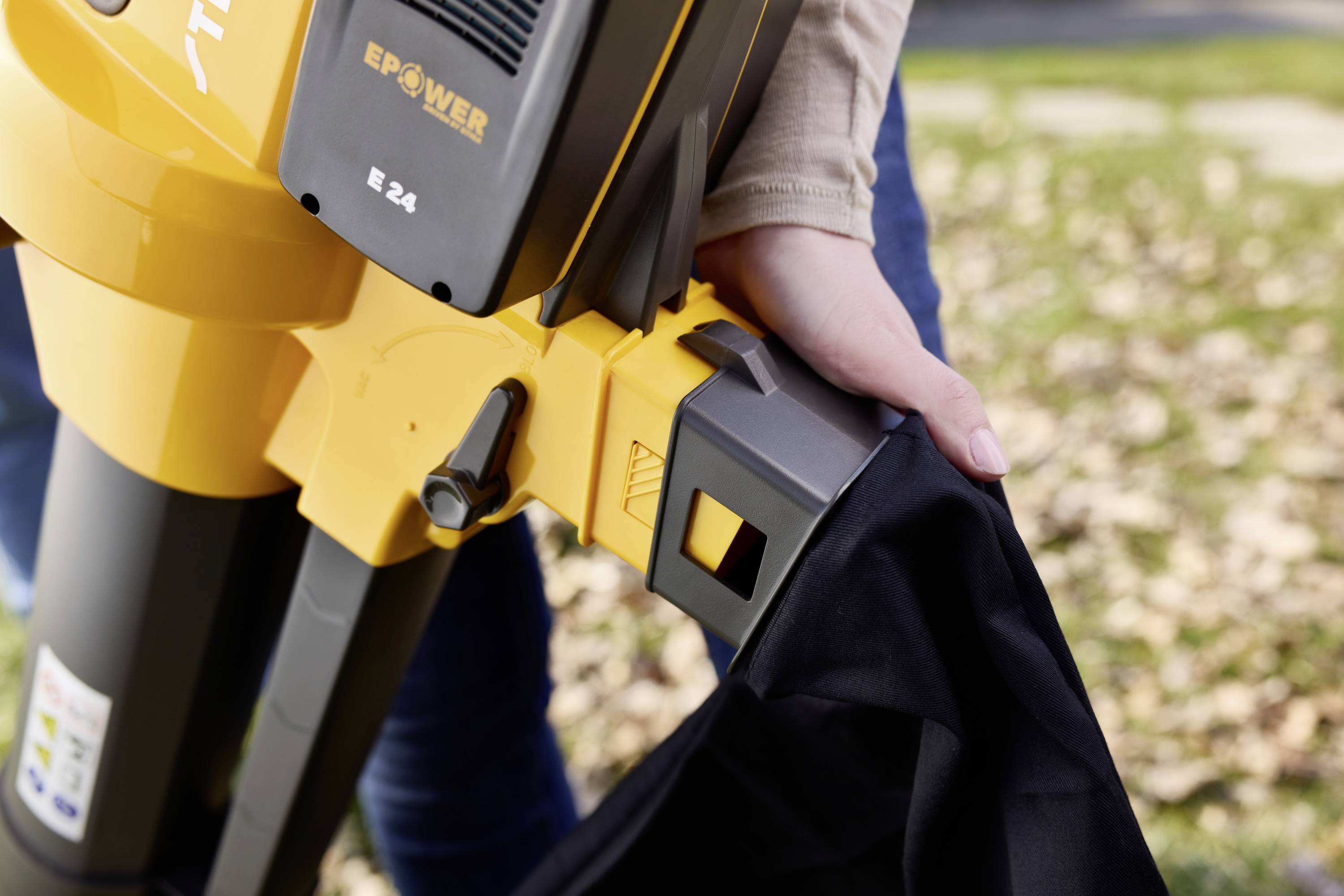 A person is attaching a black bag to a yellow leaf blower outdoors. Fallen leaves are visible in the background.
