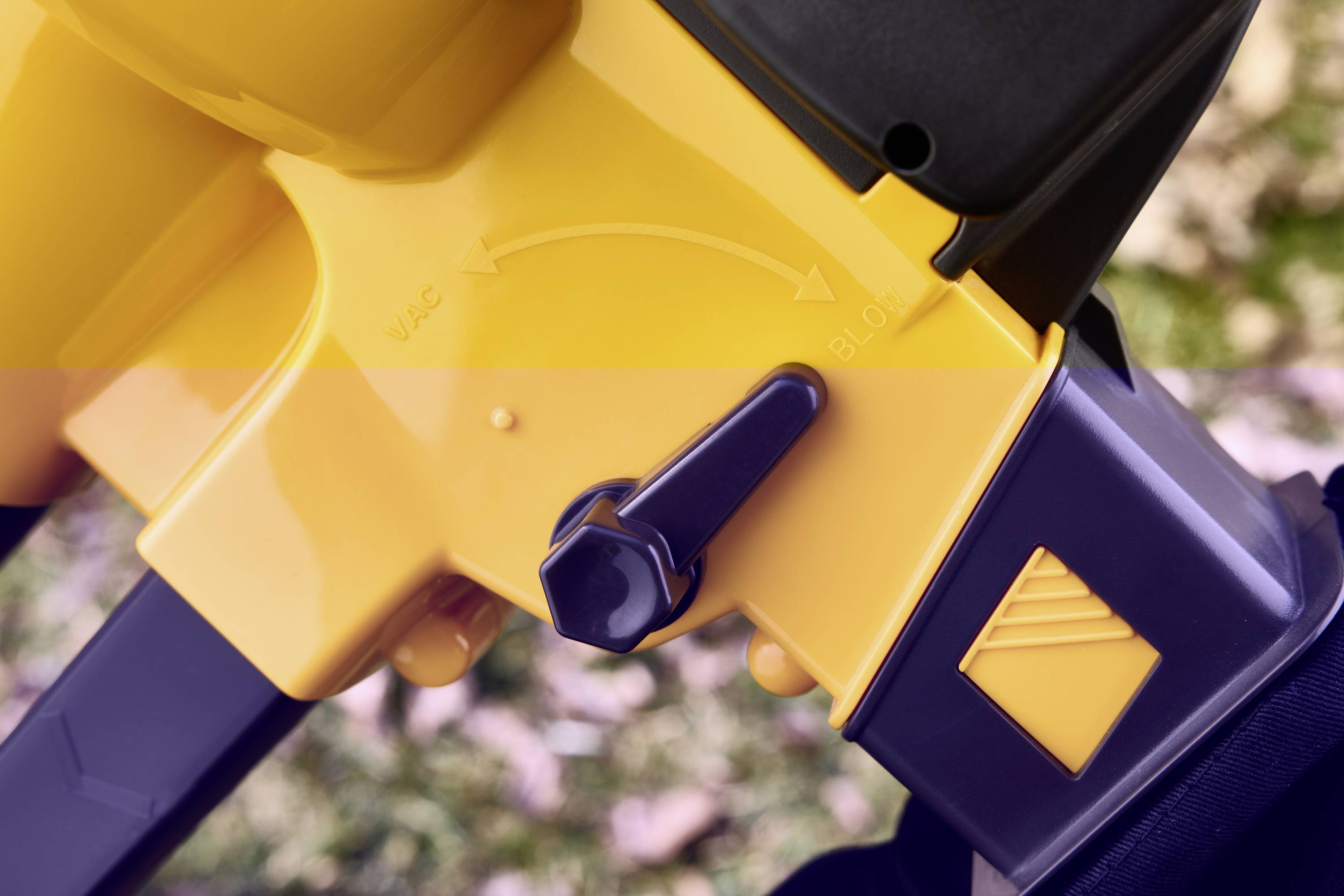 A leaf blower with a yellow casing and a switch for the 'VAC' and 'BLO' functions. Blurred leaves are visible on grass in the background.