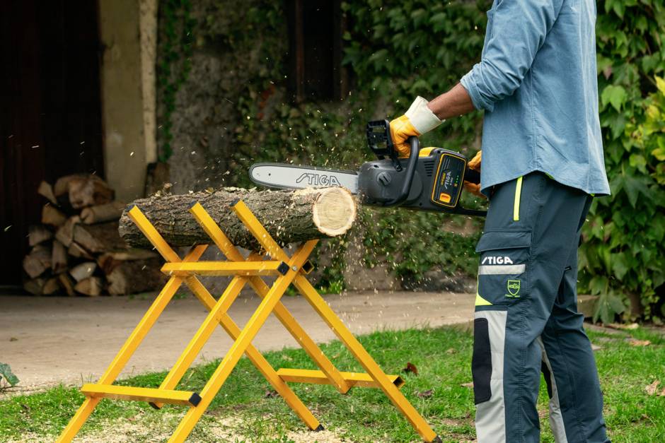 A person is cutting a wooden log with a chainsaw on a yellow wooden trestle outdoors. Stacked logs are visible in the background.