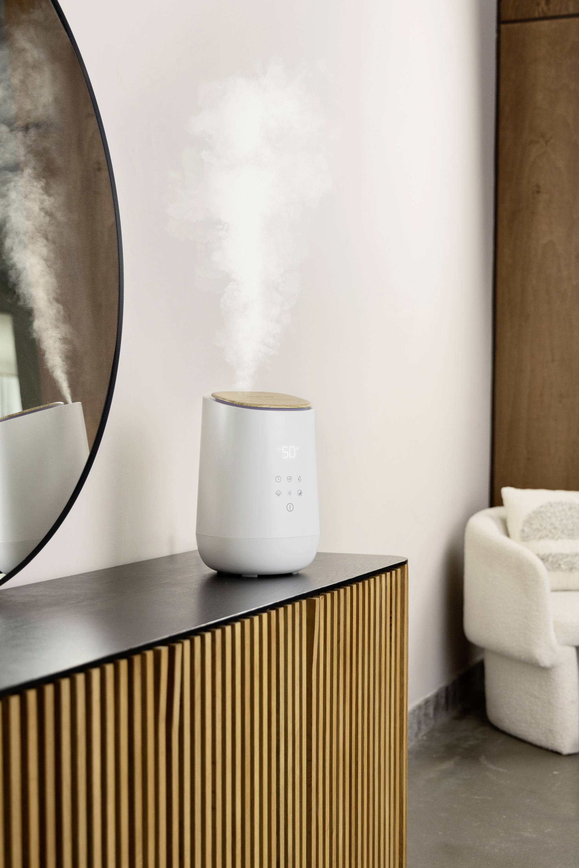 Humidifier on a wooden table, emitting steam. A mirror and an armchair in the background. Pleasant, modern room design.