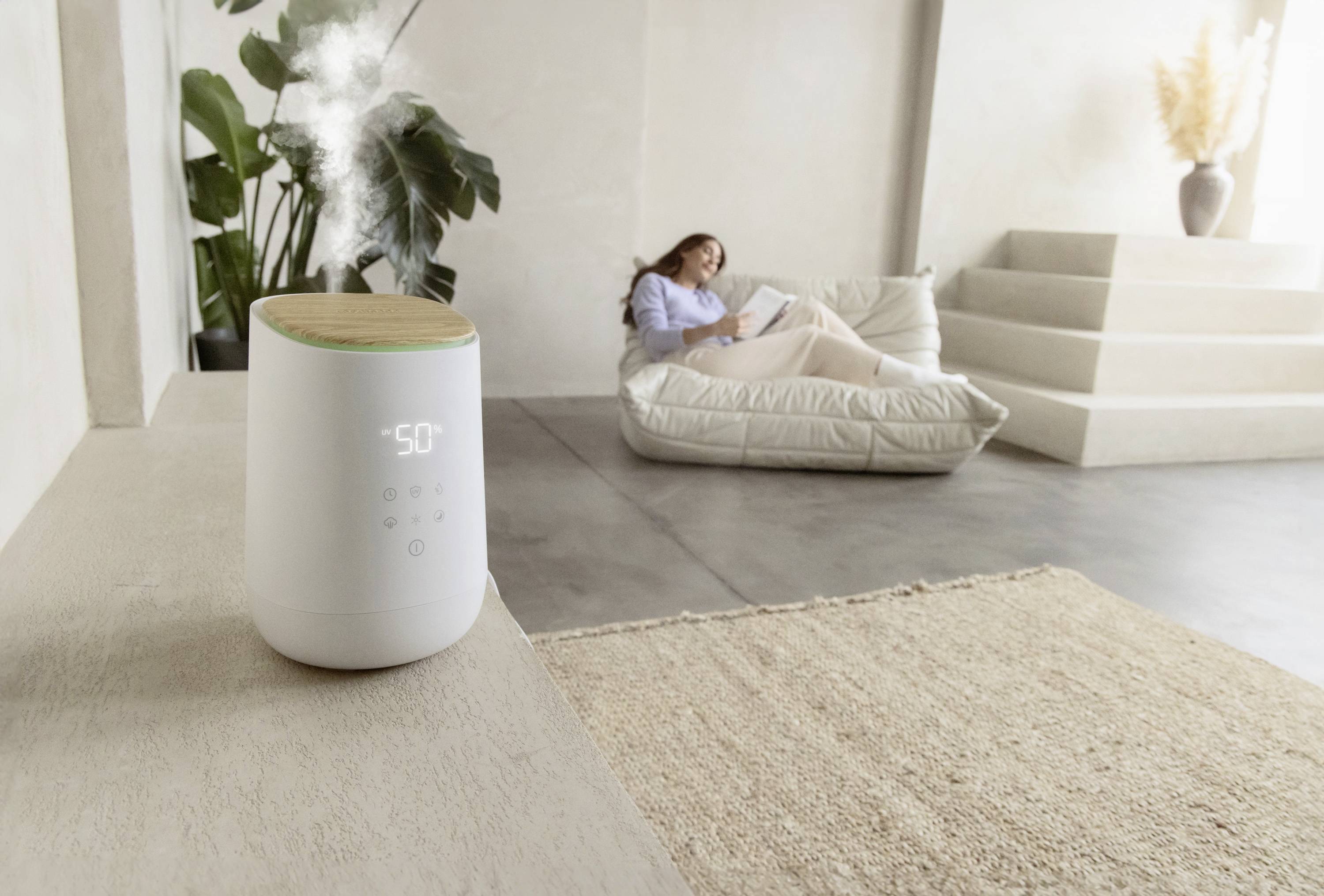 Humidifier on a table, steaming. A person relaxes on a sofa in the background. Minimalist, bright environment.