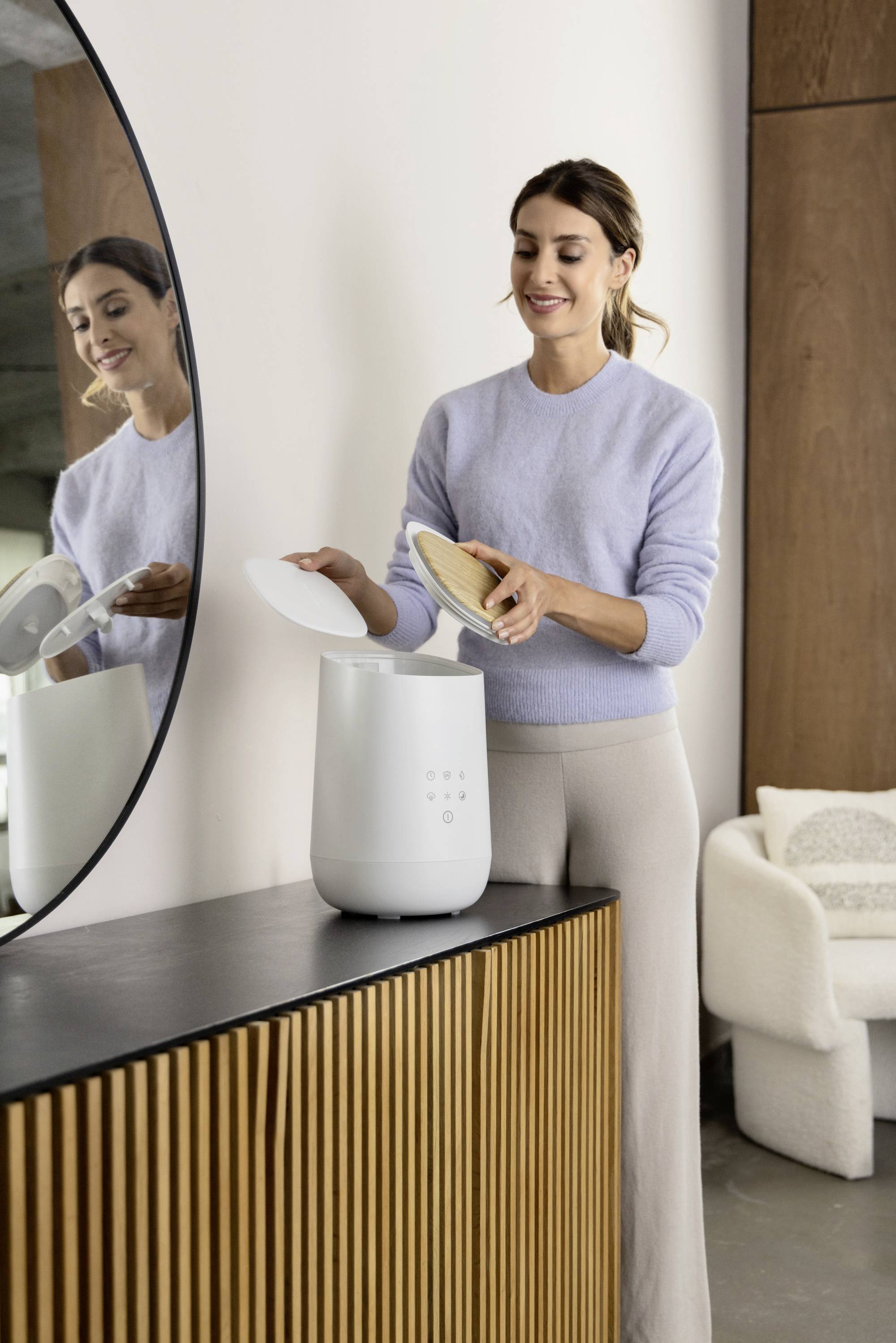A woman places bread in a white air purifier on a black table. She is wearing a light-coloured jumper and is standing in front of a mirror.