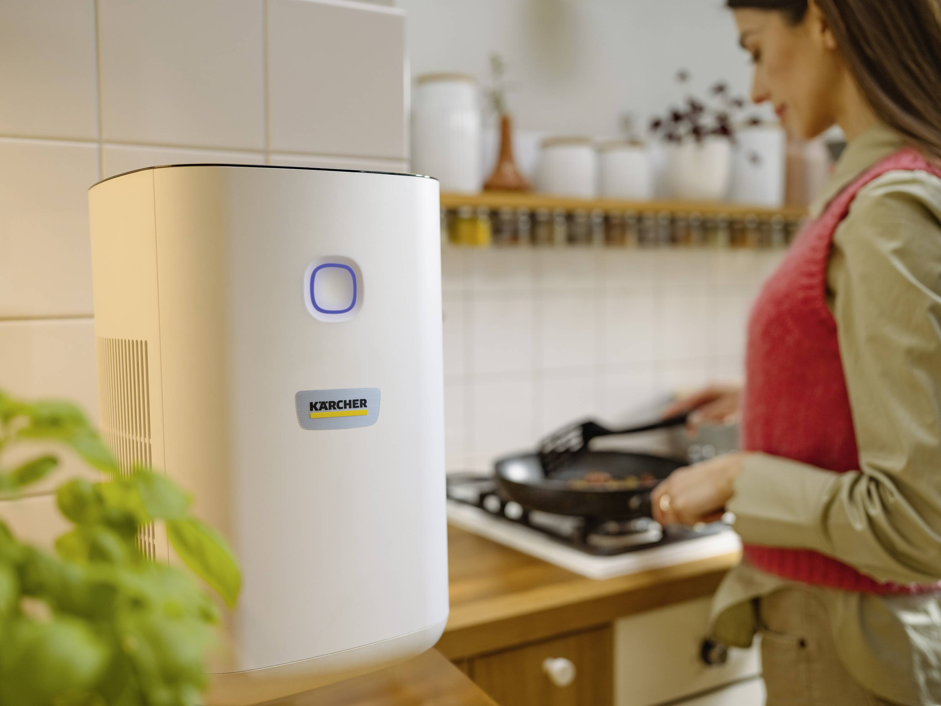 A woman is cooking in a kitchen. In the foreground, an air purifier with an illuminated control panel is visible.