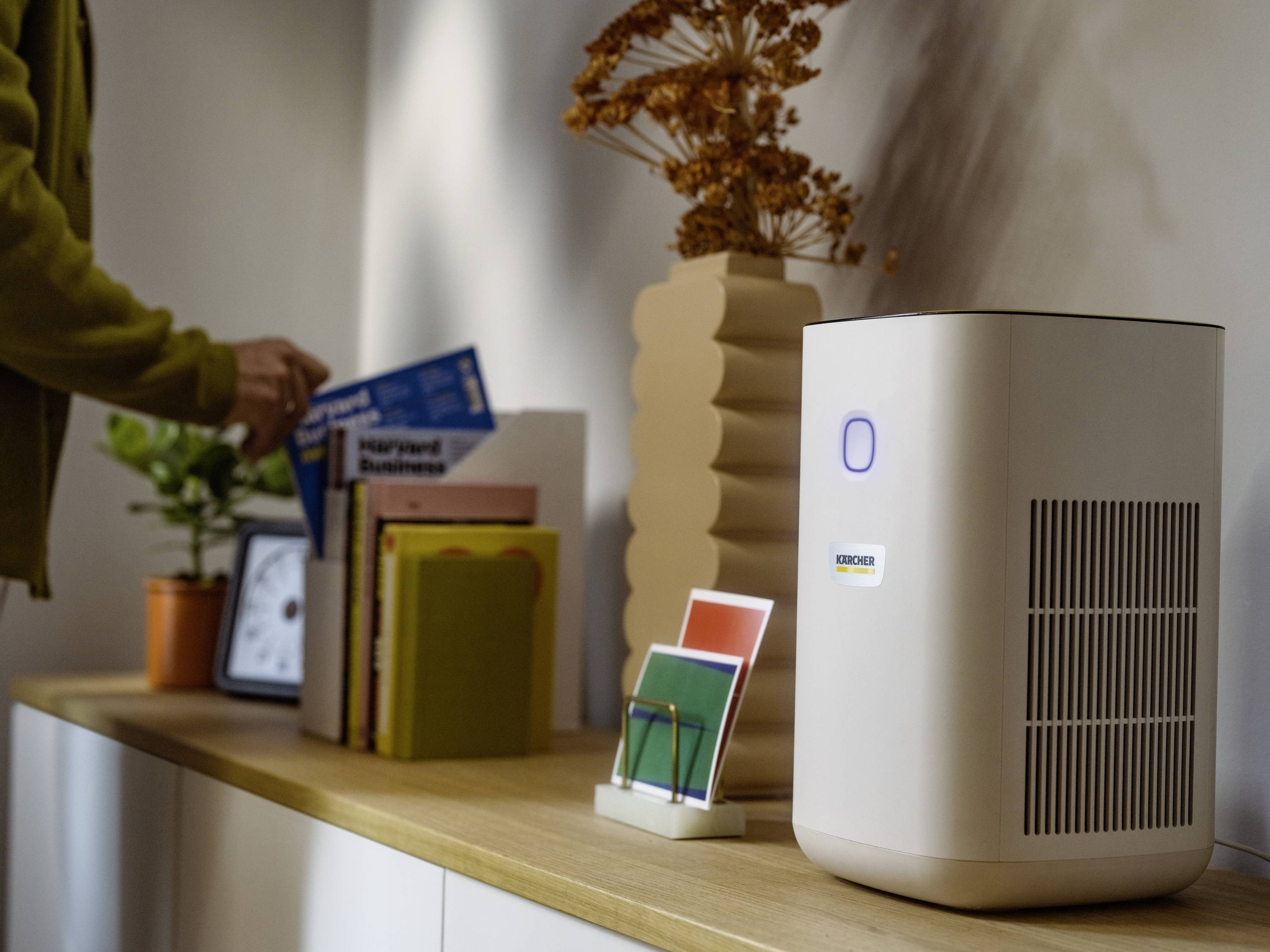 On a shelf, an air purifier sits next to books and plants. A person reaches for a magazine. Natural room decor.