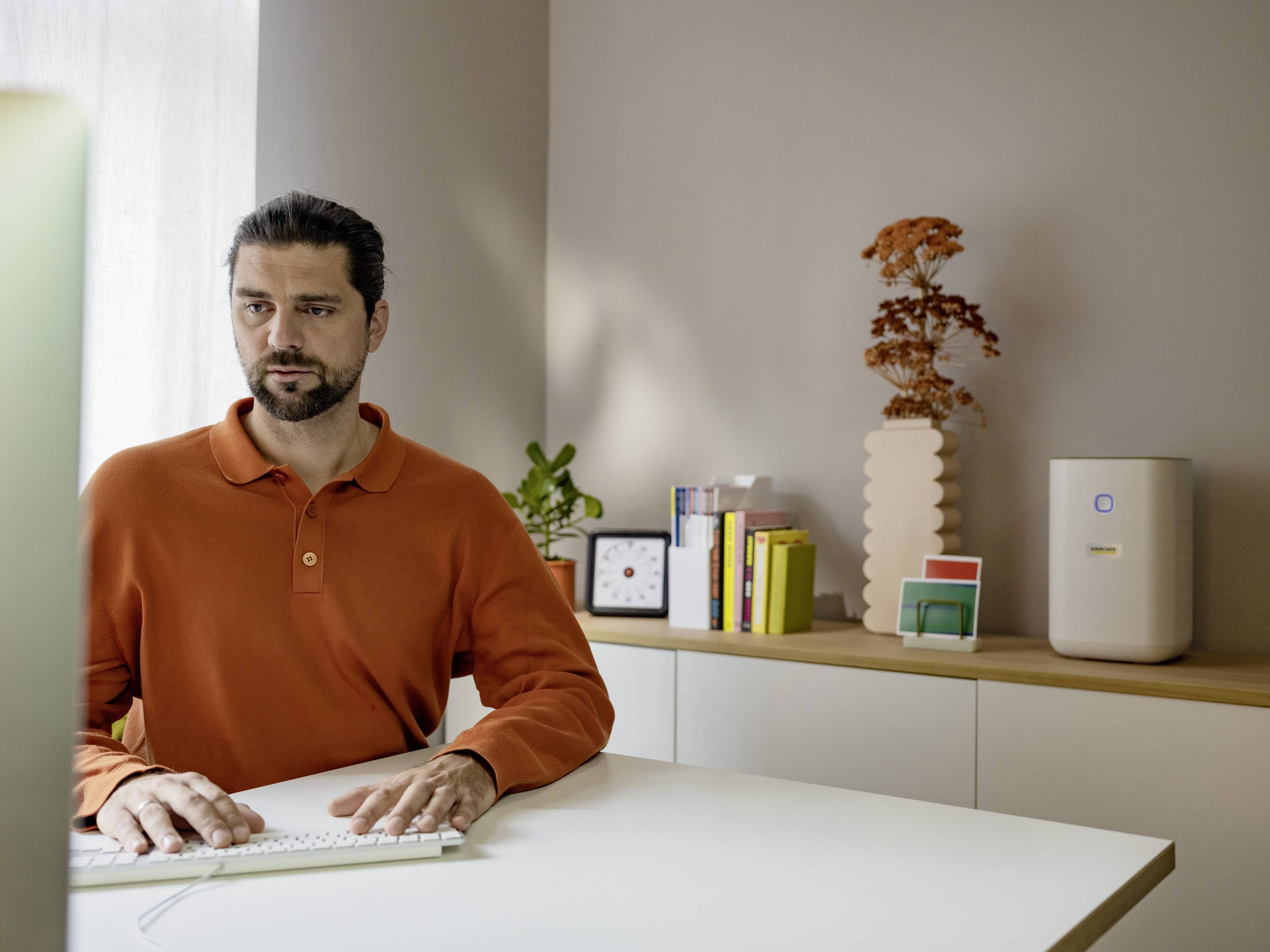 A man with a beard is typing on a keyboard in a modernly furnished office with plants and books on a shelf.