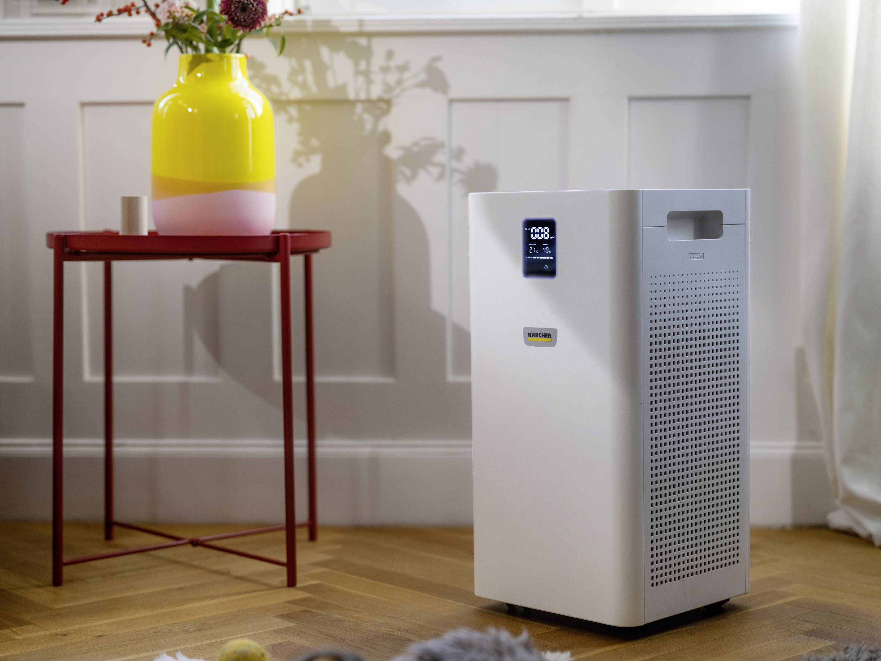 An air purifier stands on a wooden floor next to a small table with a flower-filled vase, surrounded by natural light.