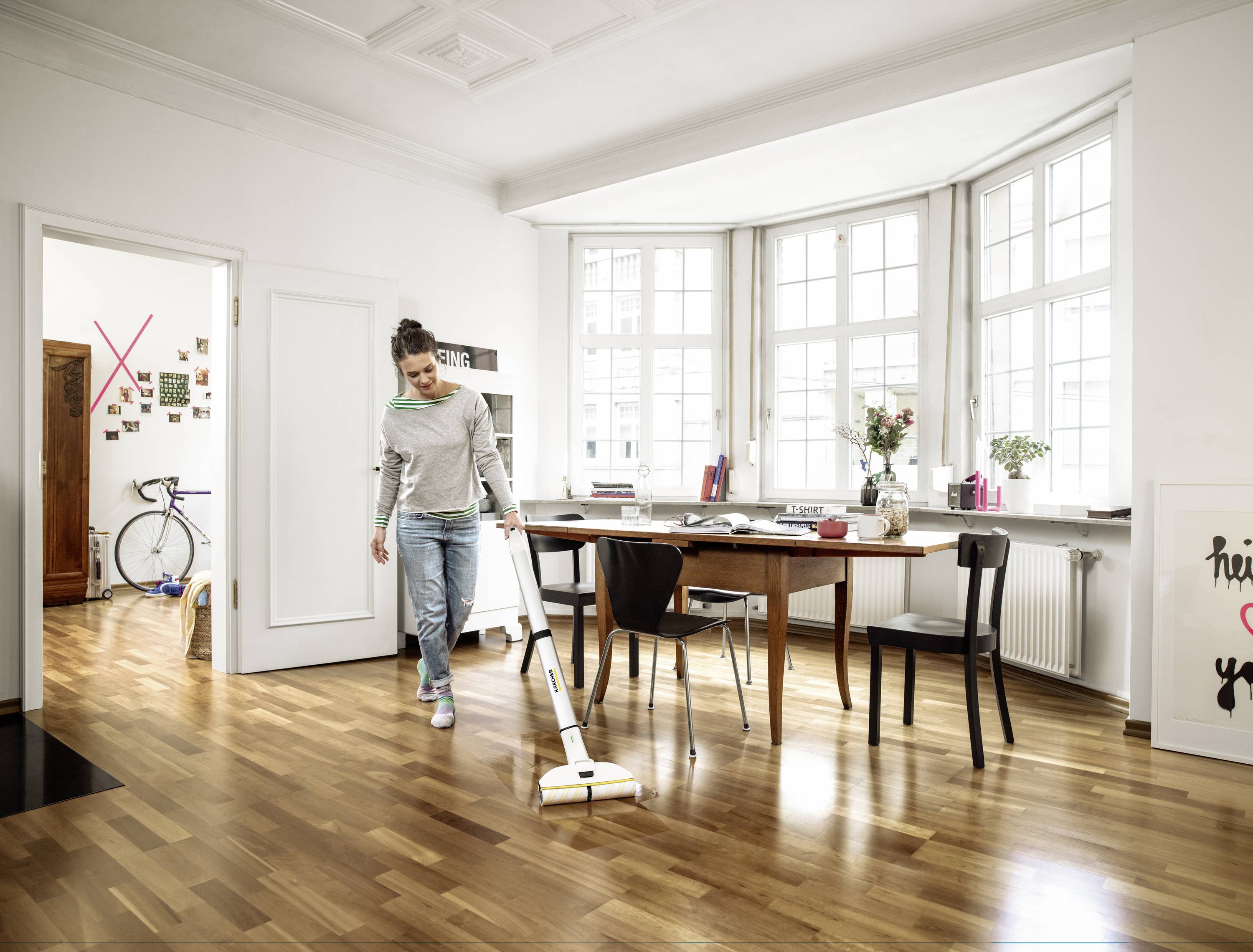 A woman is cleaning a bright, modern room with a wooden floor and a large table. In the background, there is a window, a shelf, and a bicycle.