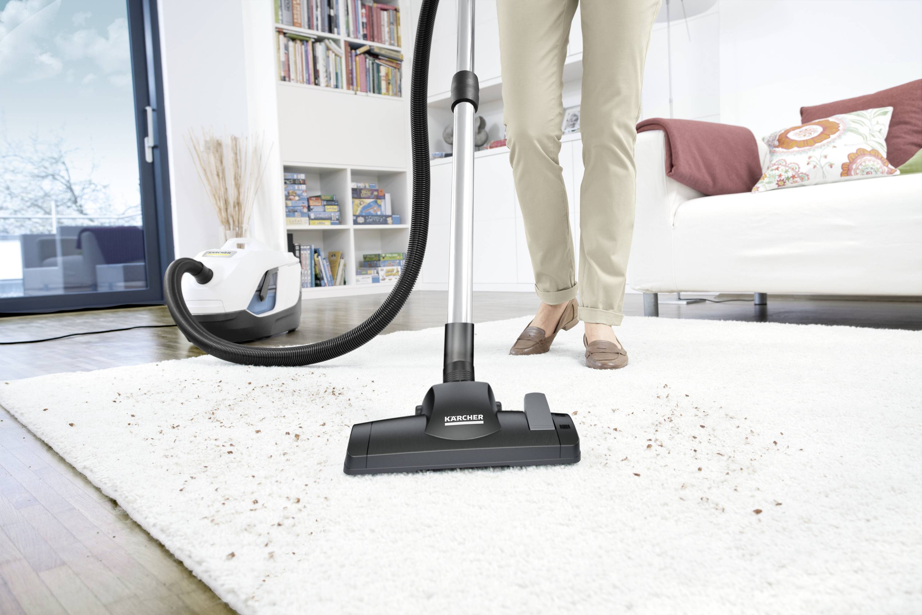 A person is vacuuming a light-coloured carpet in a living room. The hoover is removing visible dirt from the carpet. In the background, a sofa and bookshelf can be seen.