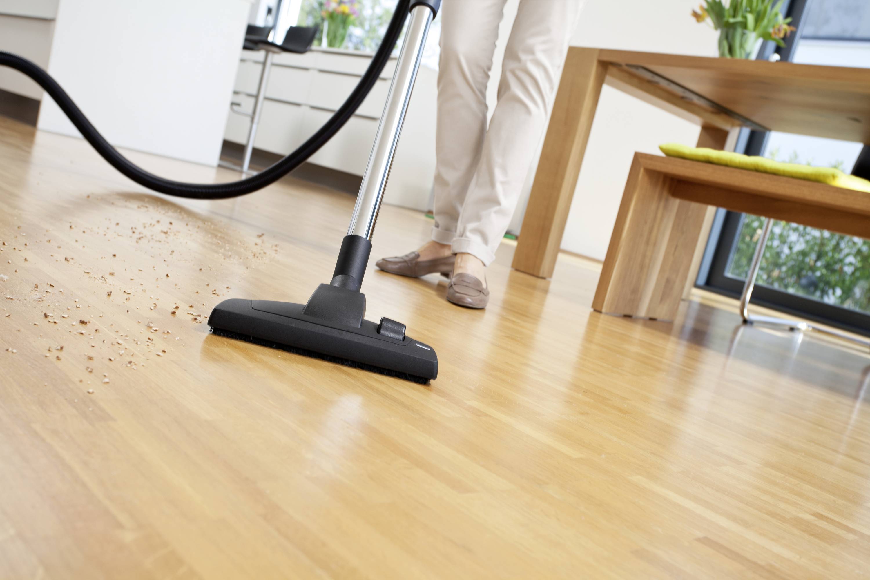 A person is hoovering a wooden floor in a bright room, removing crumbs. A table is in the background, with a window letting in daylight.