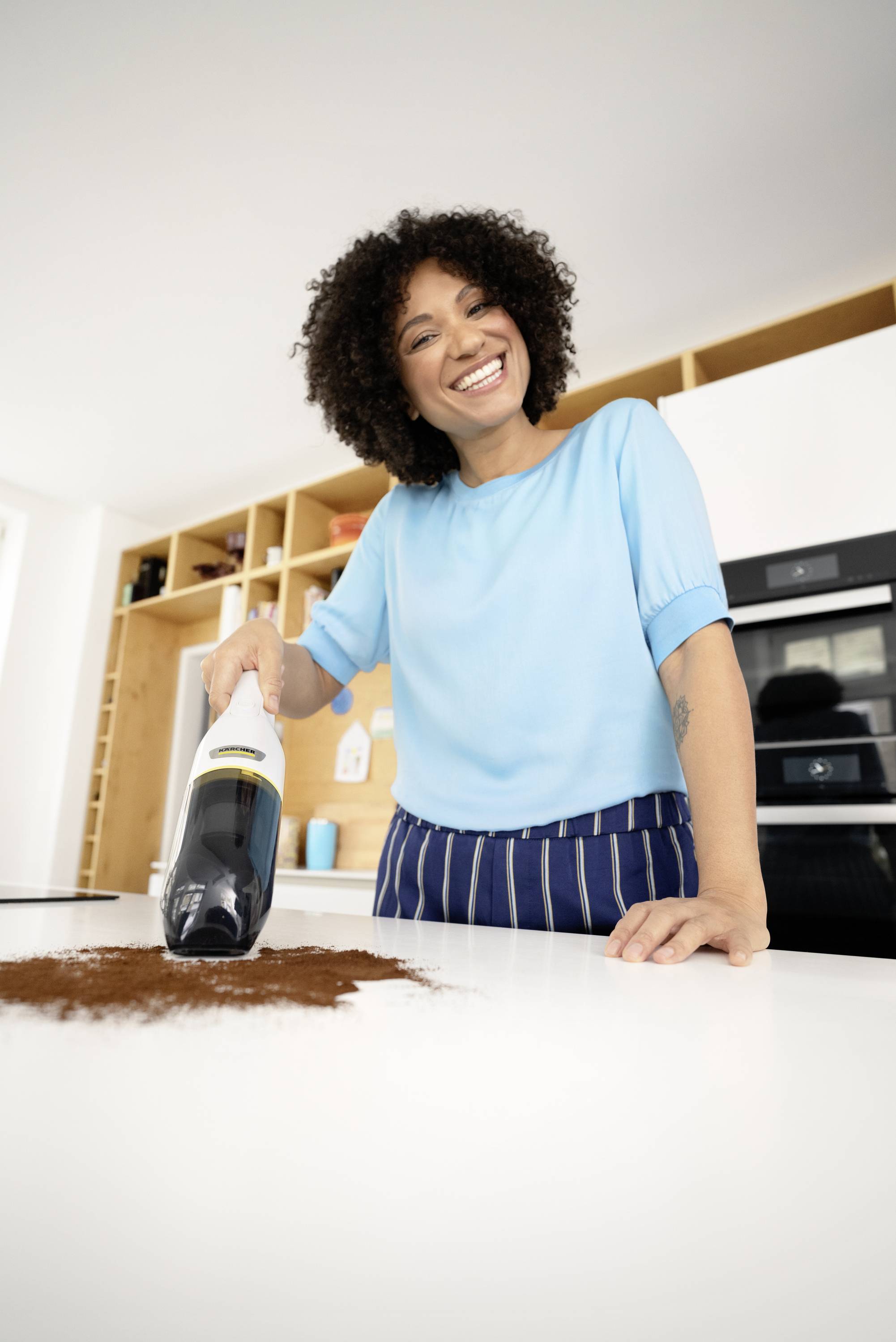 A woman smiles as she uses a handheld vacuum cleaner to remove coffee grounds from a white kitchen worktop. Kitchen shelves and a cooker are visible in the background.