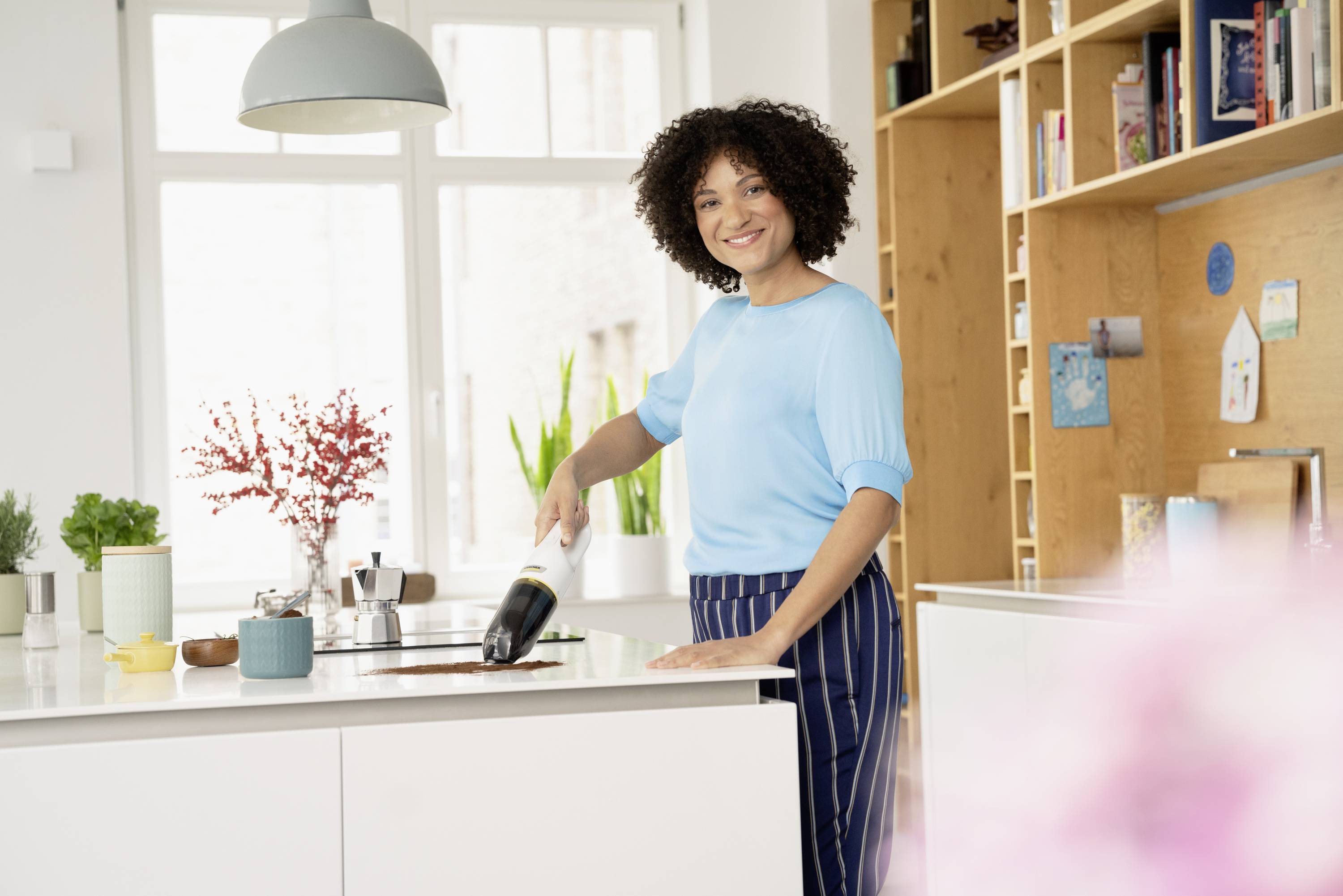 A smiling woman in a kitchen is using a handheld vacuum to clean up crumbs from a worktop. In the background, plants and shelves are visible.