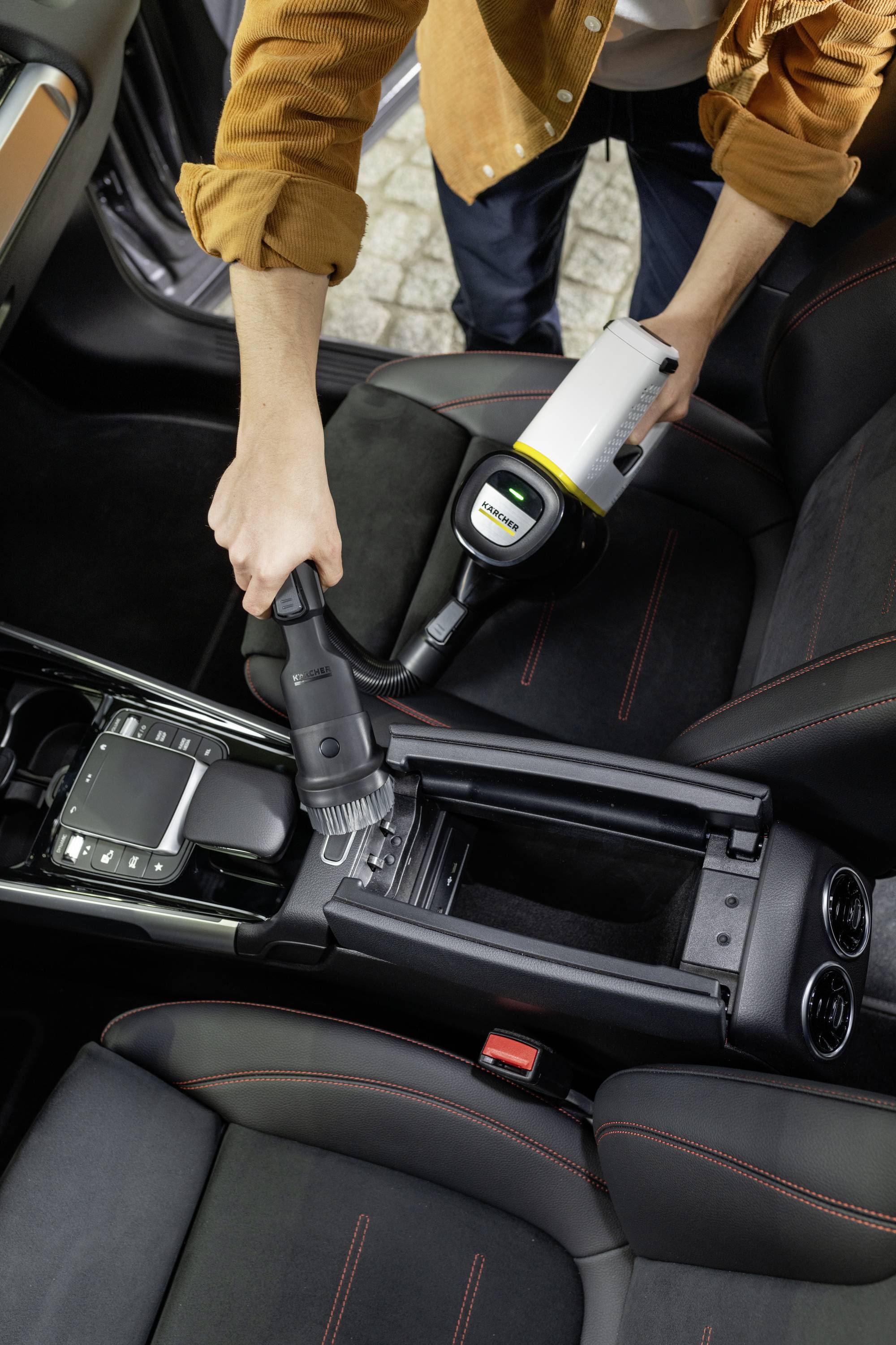A person is vacuuming a car interior with a portable hoover. Focus is on cleaning the centre console between the front seats.
