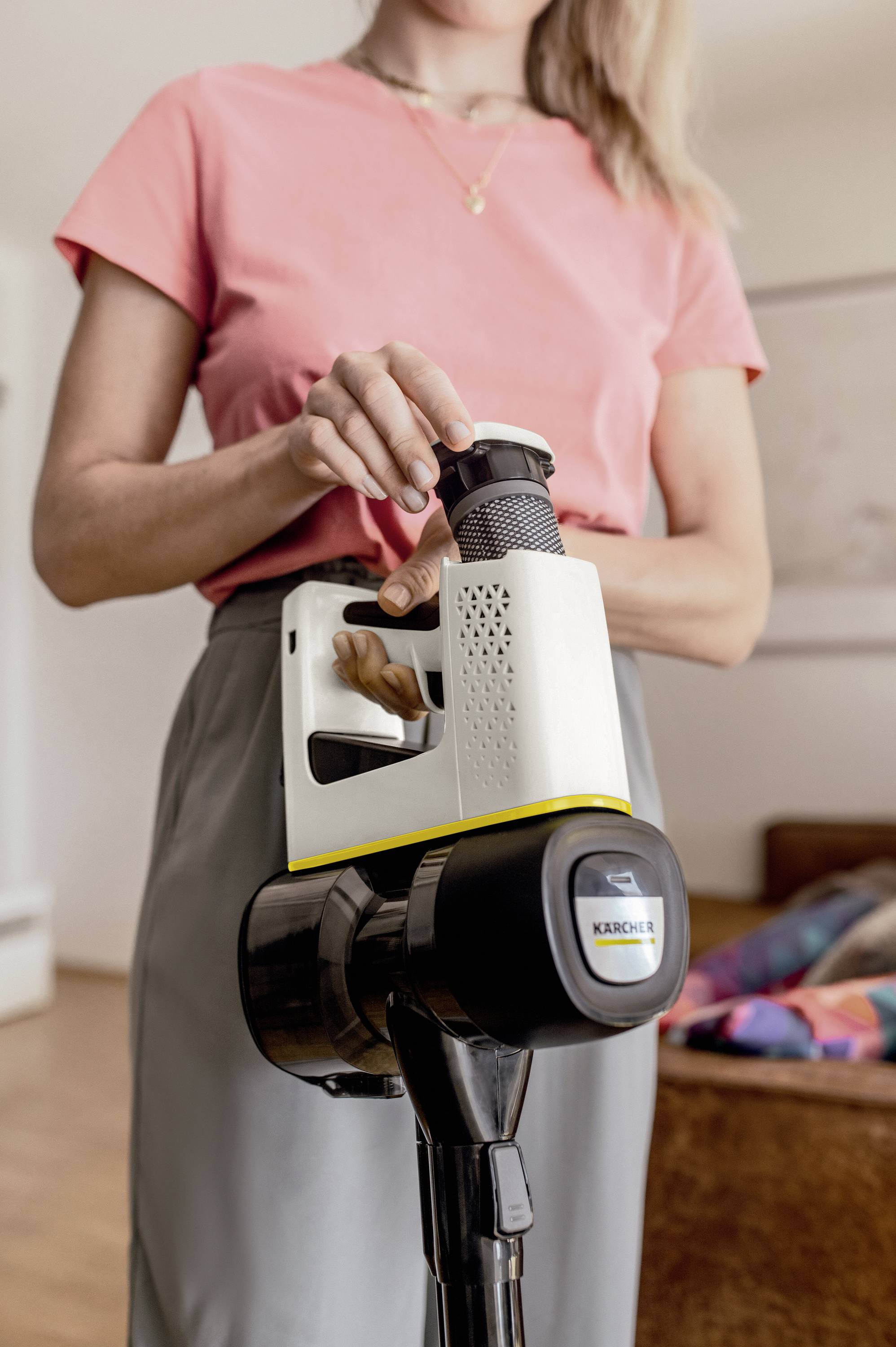 A person in a pink T-shirt is holding a cordless vacuum cleaner in a living room. The vacuum cleaner has a yellow and black design.
