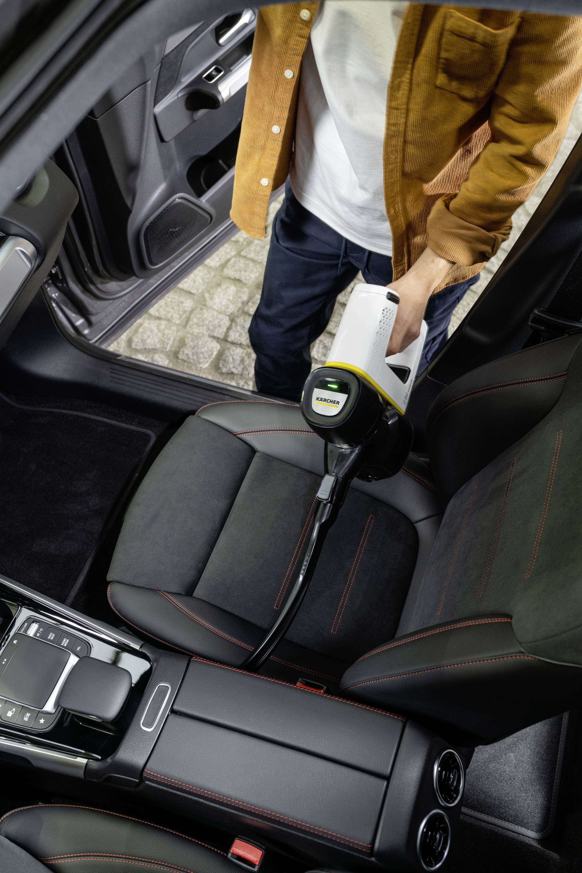 A person is using a handheld vacuum cleaner to clean the passenger seat of a car. The door is open, and a stone floor surface can be seen.