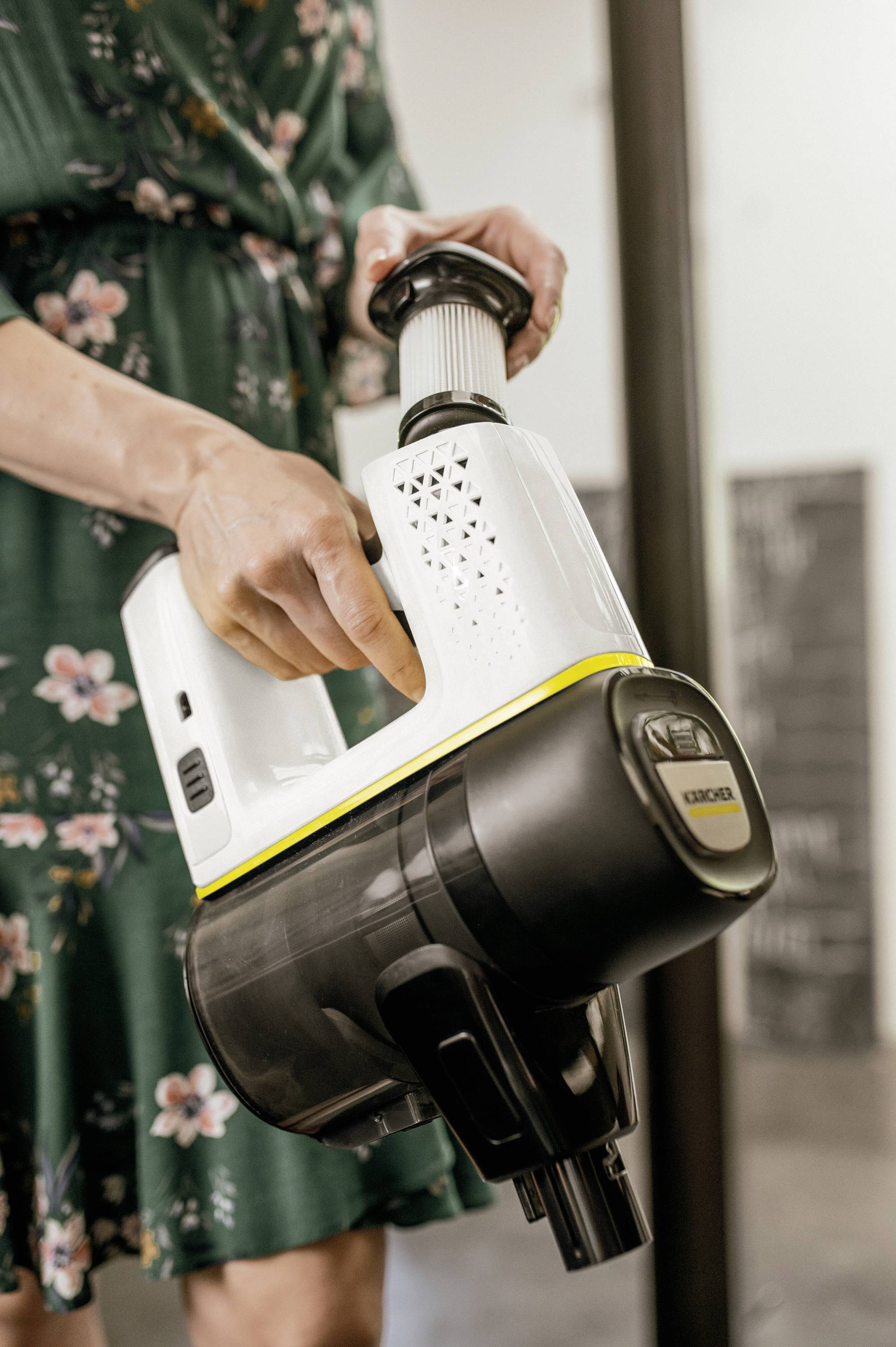 A person is holding a white and black cordless vacuum cleaner. They are wearing a green dress with a floral pattern.